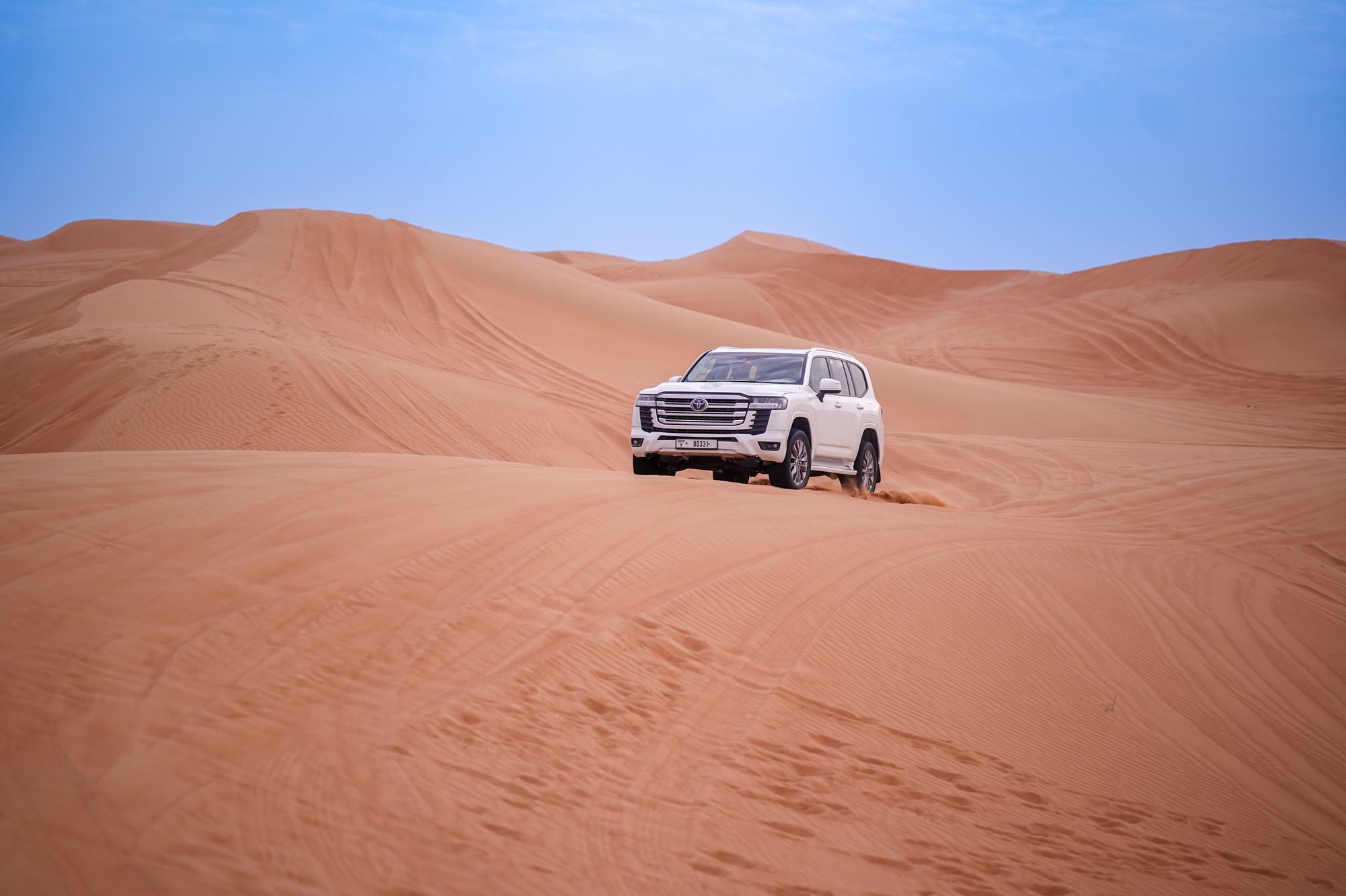 A white 300 Series Land Cruiser driving across red sand dunes under a pale blue sky in the UAE desert