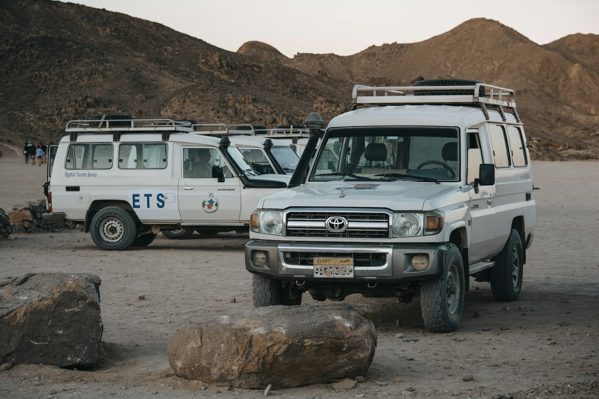 White Land Cruiser 70 Series troop carriers with roof racks parked among boulders in a rocky desert valley