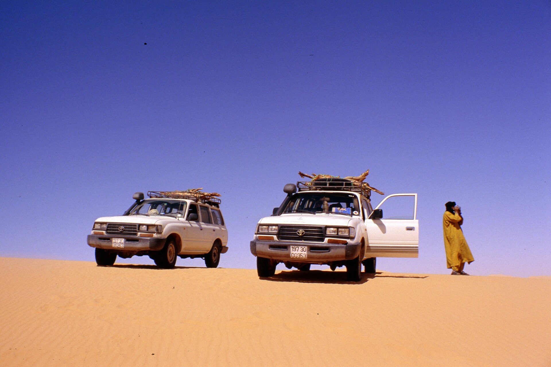 Two white post-1995 Land Cruiser 80 Series wagons with snorkels and loaded roof racks on a sand dune crest in the Algerian Sahara
