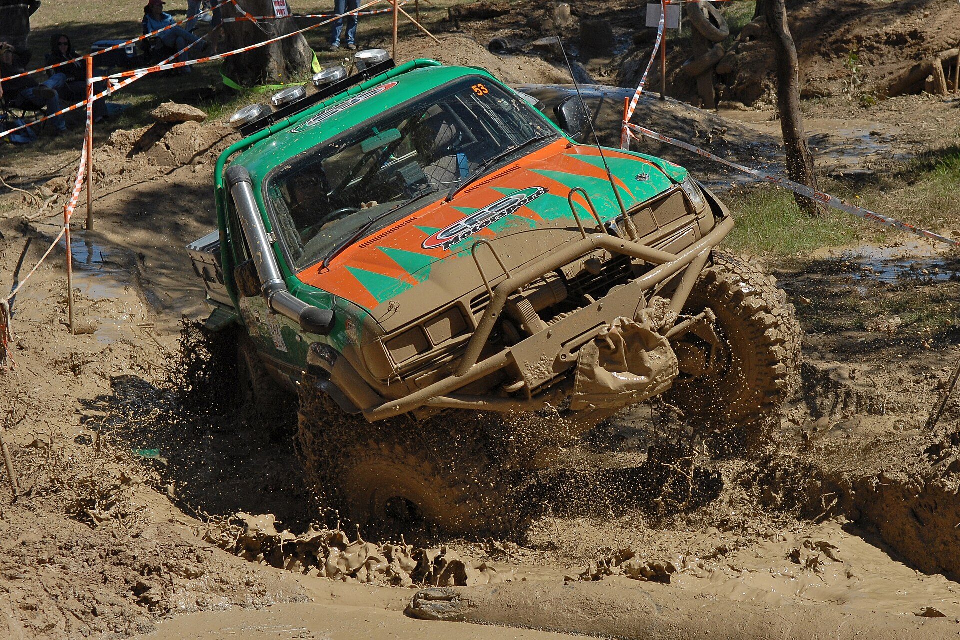 A green and orange competition Land Cruiser 80 Series, cut down to a cab-and-tray body with a snorkel and bull bar, throwing mud as it climbs out of a deep rutted bog