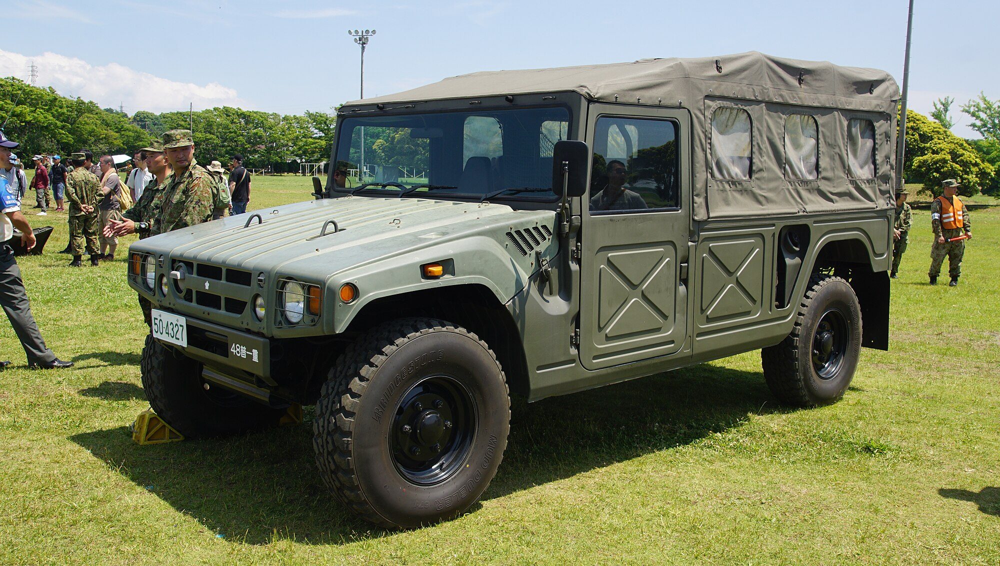 An olive-green JGSDF High Mobility Vehicle, the military BXD10-family version of the Mega Cruiser, with a canvas top parked on grass at a base open day