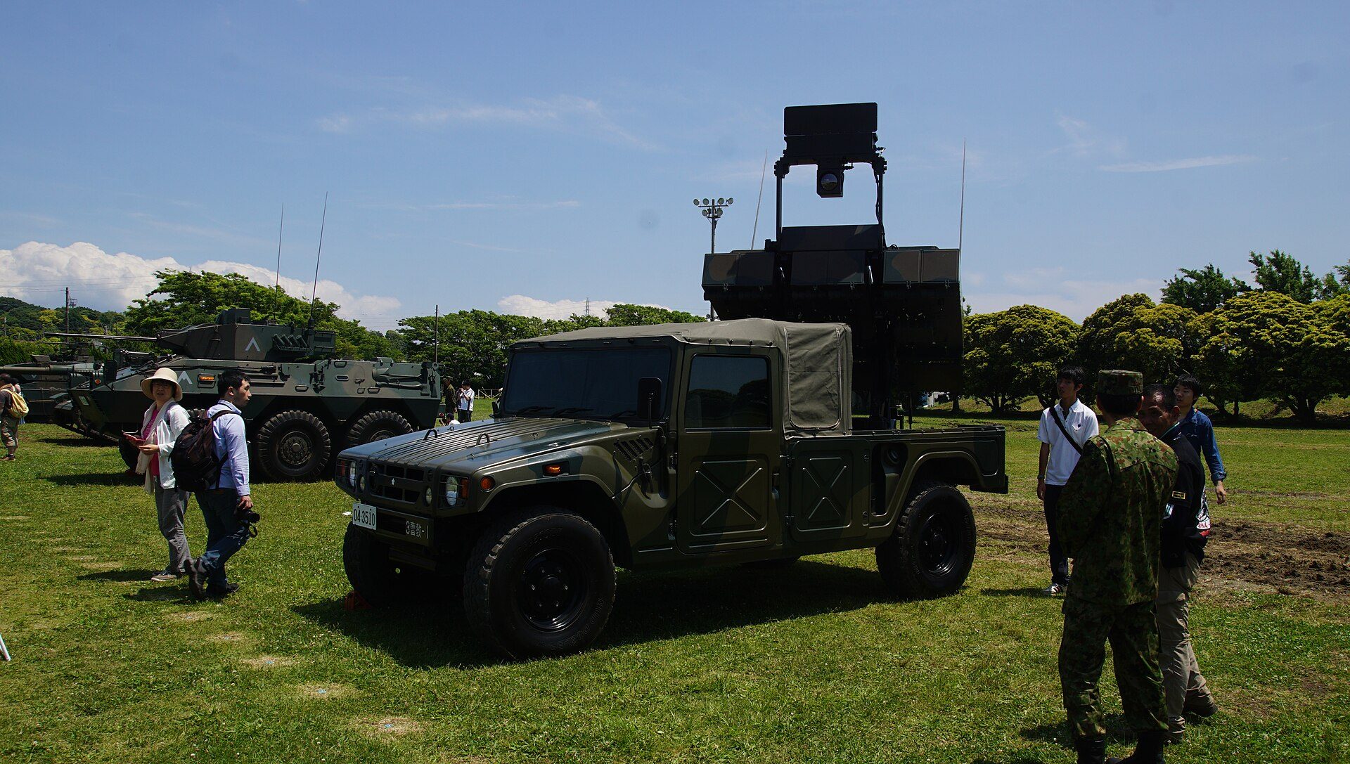 A JGSDF High Mobility Vehicle carrying an anti-tank missile launcher on its rear deck, parked on grass among visitors