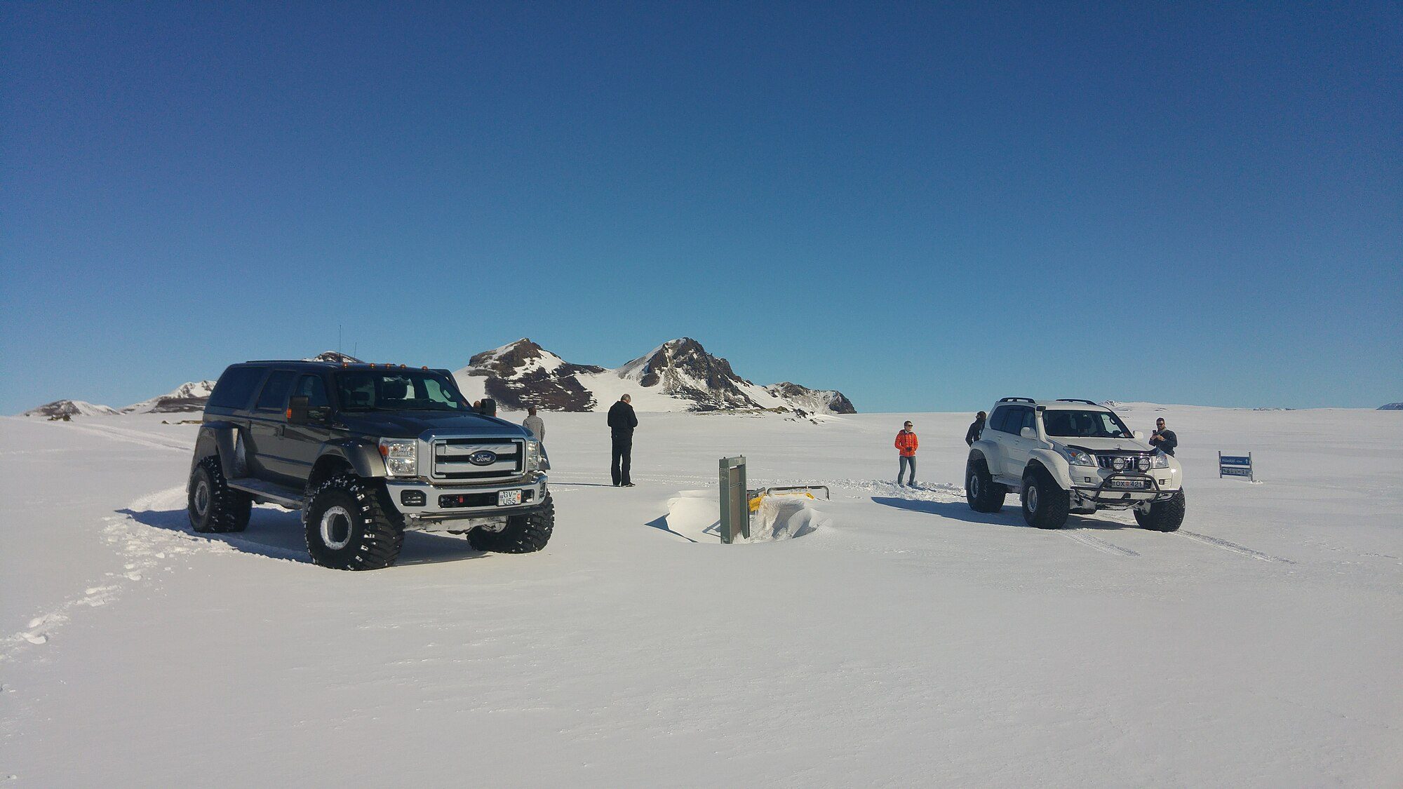 A white Land Cruiser Prado 120 'super jeep' on huge tires standing on an Icelandic snowfield next to a modified Ford