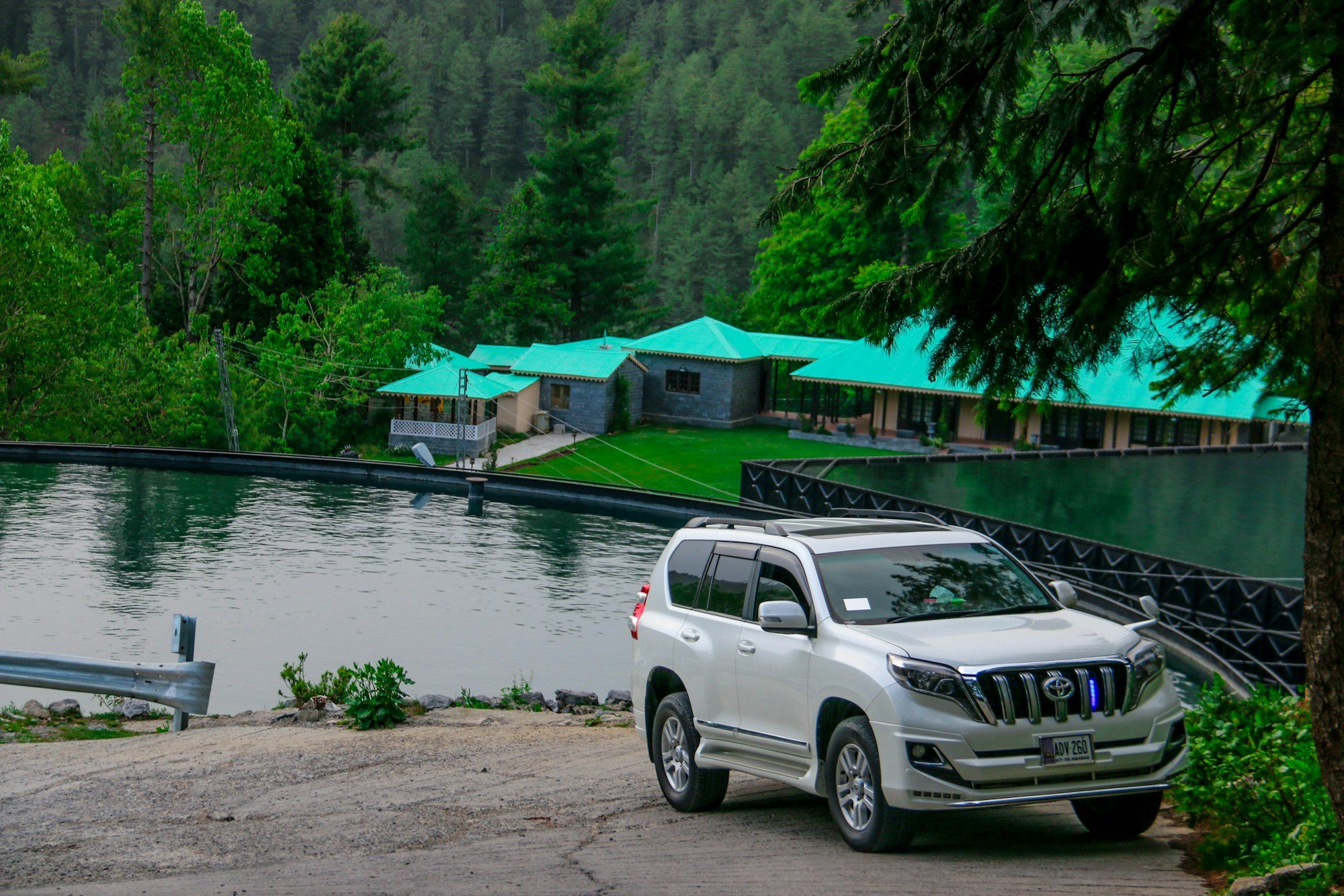 A white 2017-or-later Prado 150 parked on a gravel verge beside a lake in the pine-covered hills of Dunga Gali, Pakistan