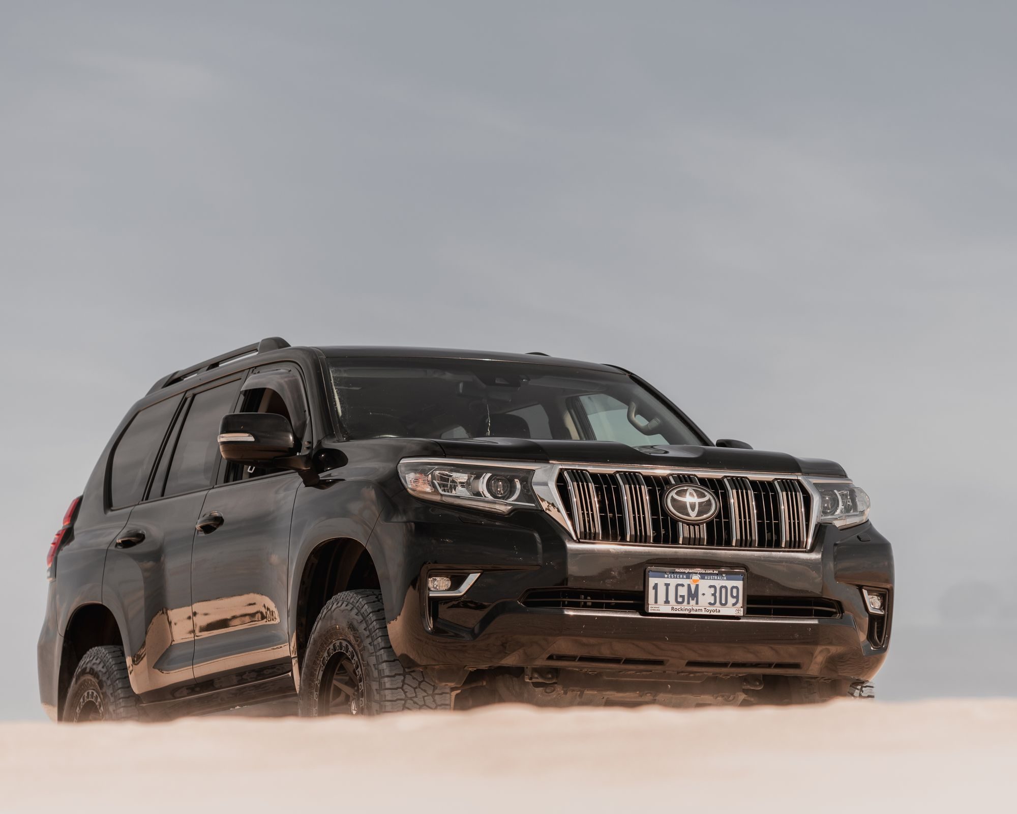 Front three-quarter view of a black 2017-or-later Prado 150 on the sand at Lancelin, showing the vertical-bar grille of the second facelift
