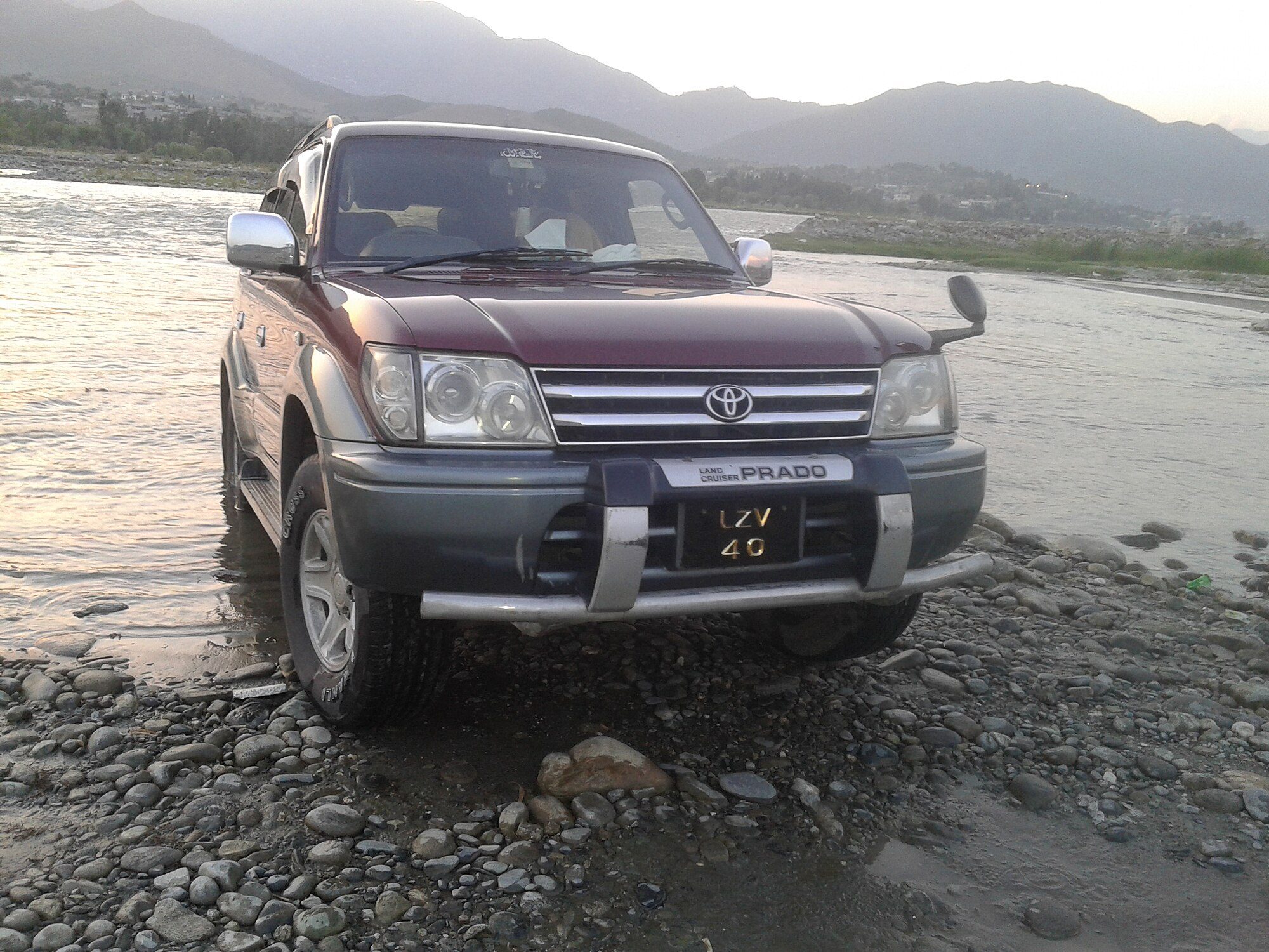 A maroon and grey Land Cruiser Prado 90 TX parked on a stony riverbed with mountains behind
