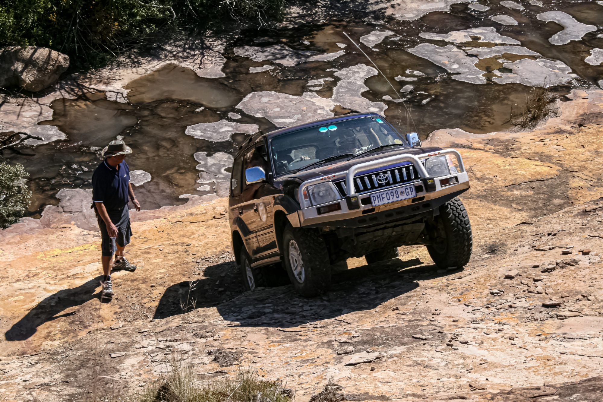 A dark facelifted Land Cruiser Prado 90 with a bull bar climbing a sandstone rock slab while a spotter walks alongside