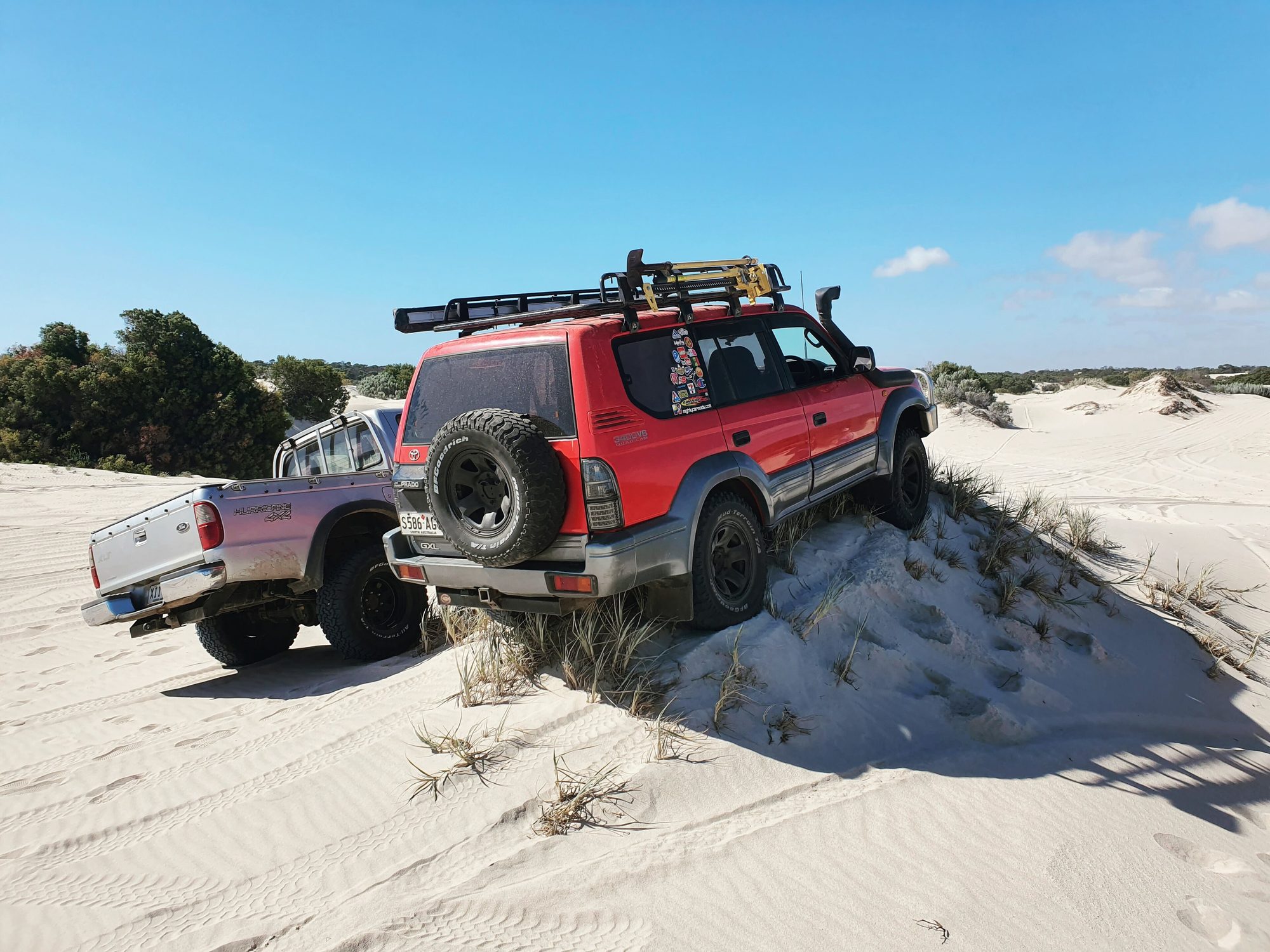 A red Land Cruiser Prado 90 GXL five-door with a roof rack, snorkel and high-lift jack perched on a grassy sand dune beside a pickup