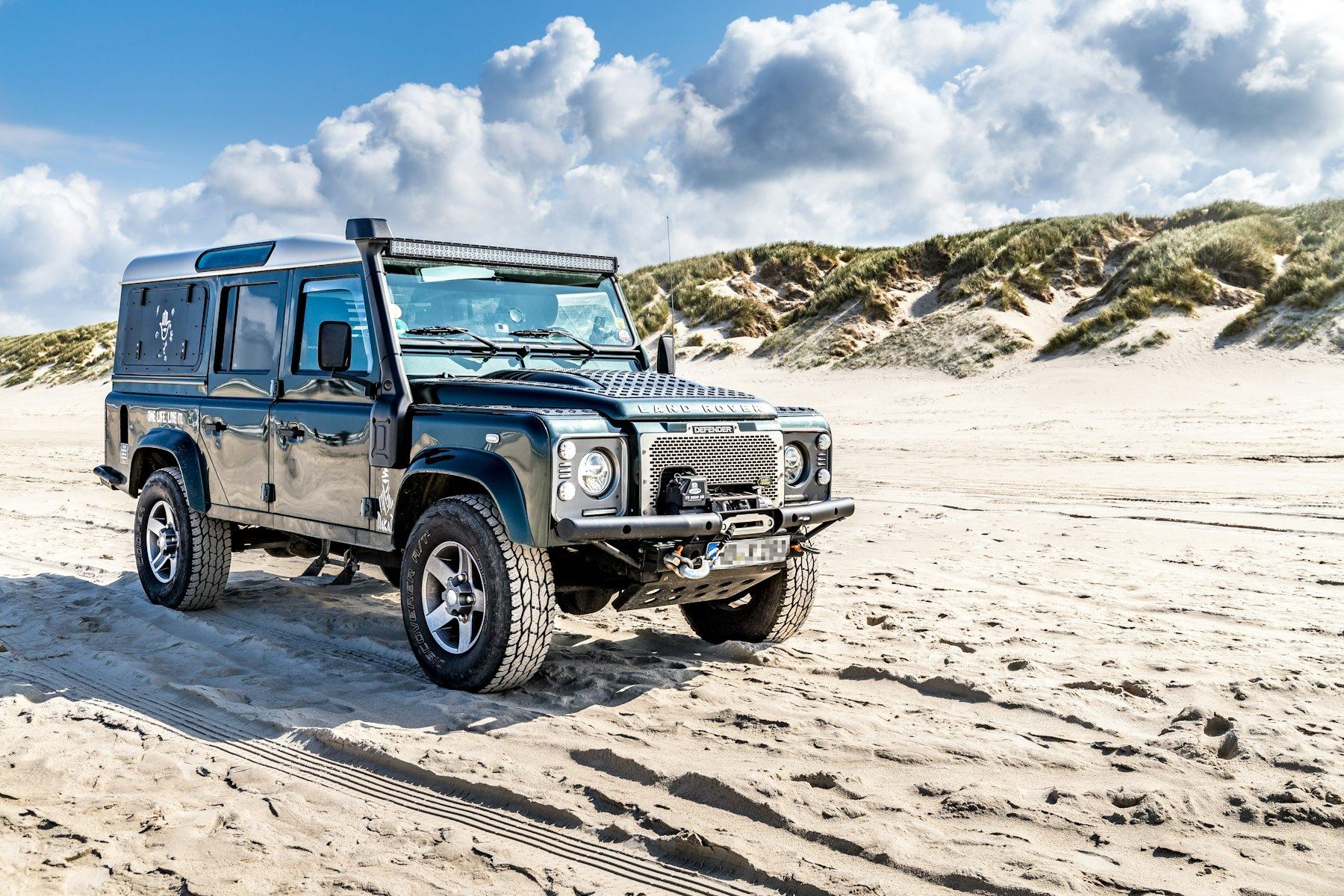A dark green classic Defender 110 with light bar and winch bumper on white sand dunes at Vejers Strand, Denmark