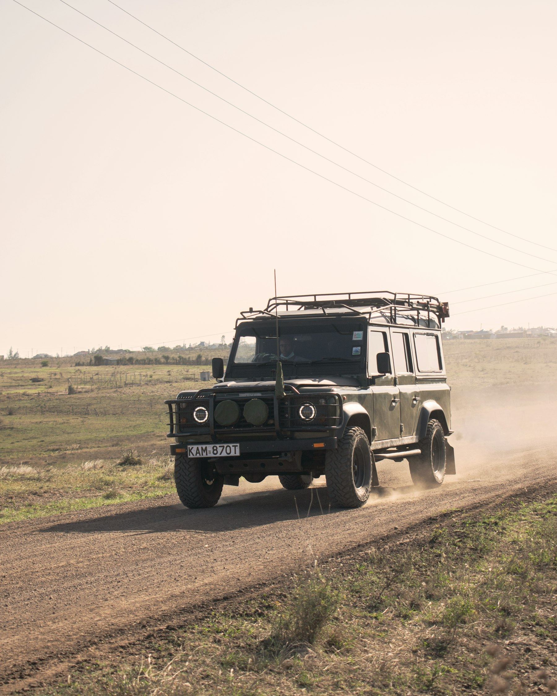 A dark classic Defender 110 with roof rack kicking up dust on a dirt road across open grassland