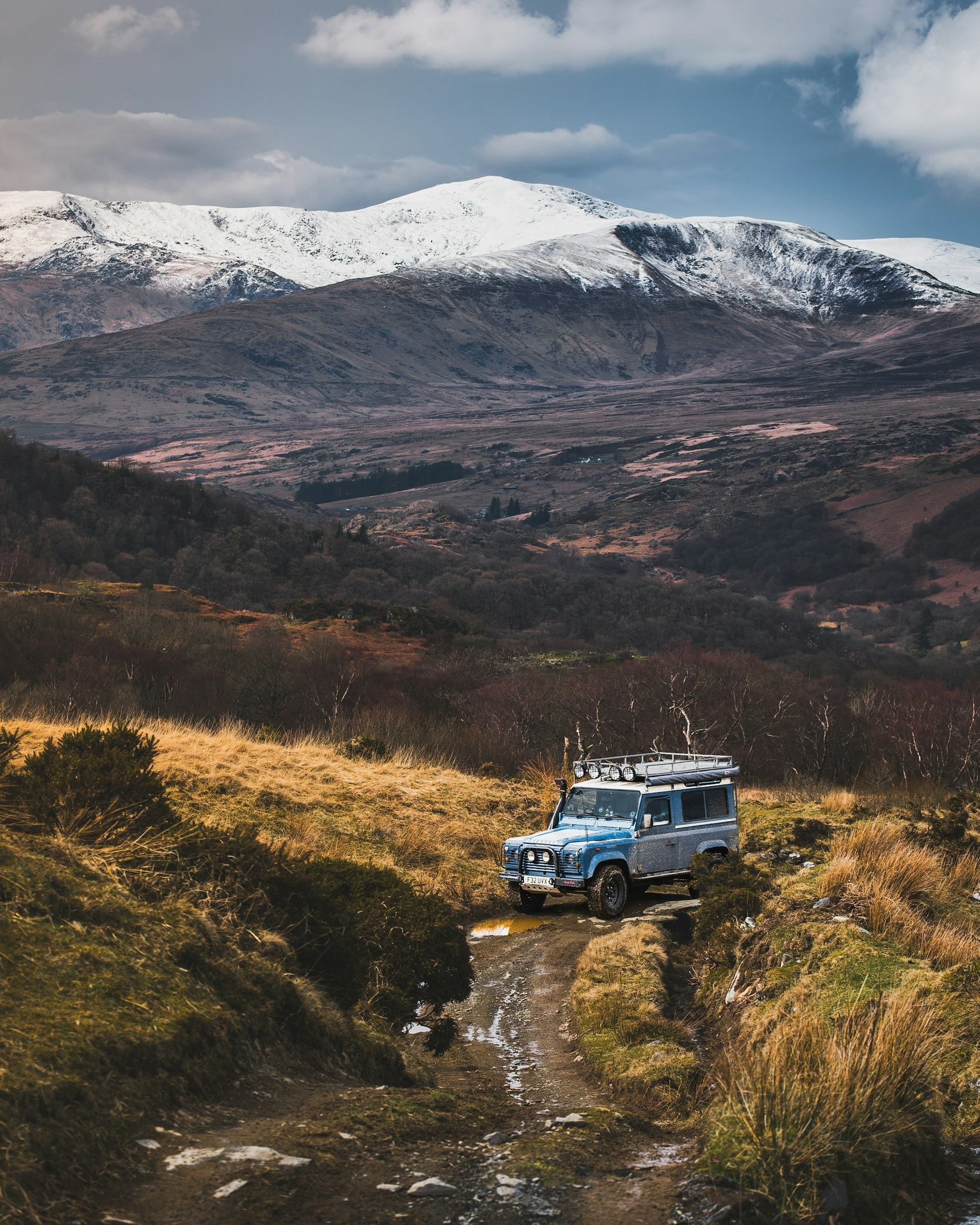 A blue classic Defender 90 with snorkel, roof rack and spotlights on a muddy moorland track below snow-capped mountains