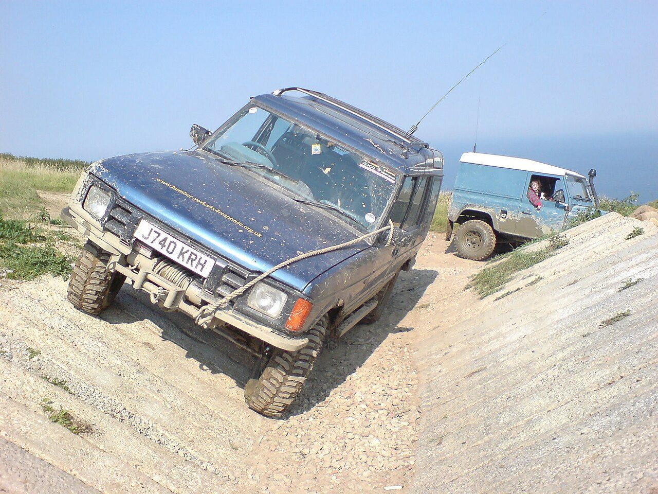 A muddy blue pre-facelift Discovery Series I with a winch bumper and mud tyres tilted in a concrete-sided off-road trench, a Defender hard-top waiting behind