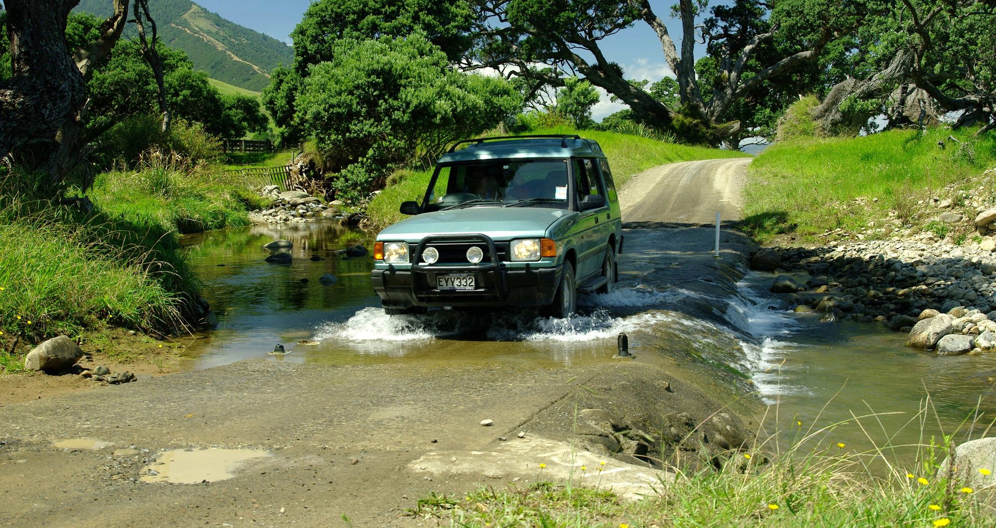 A pale-green facelifted Land Rover Discovery Series I with a nudge bar splashing through a shallow ford on a gravel road, Coromandel, New Zealand
