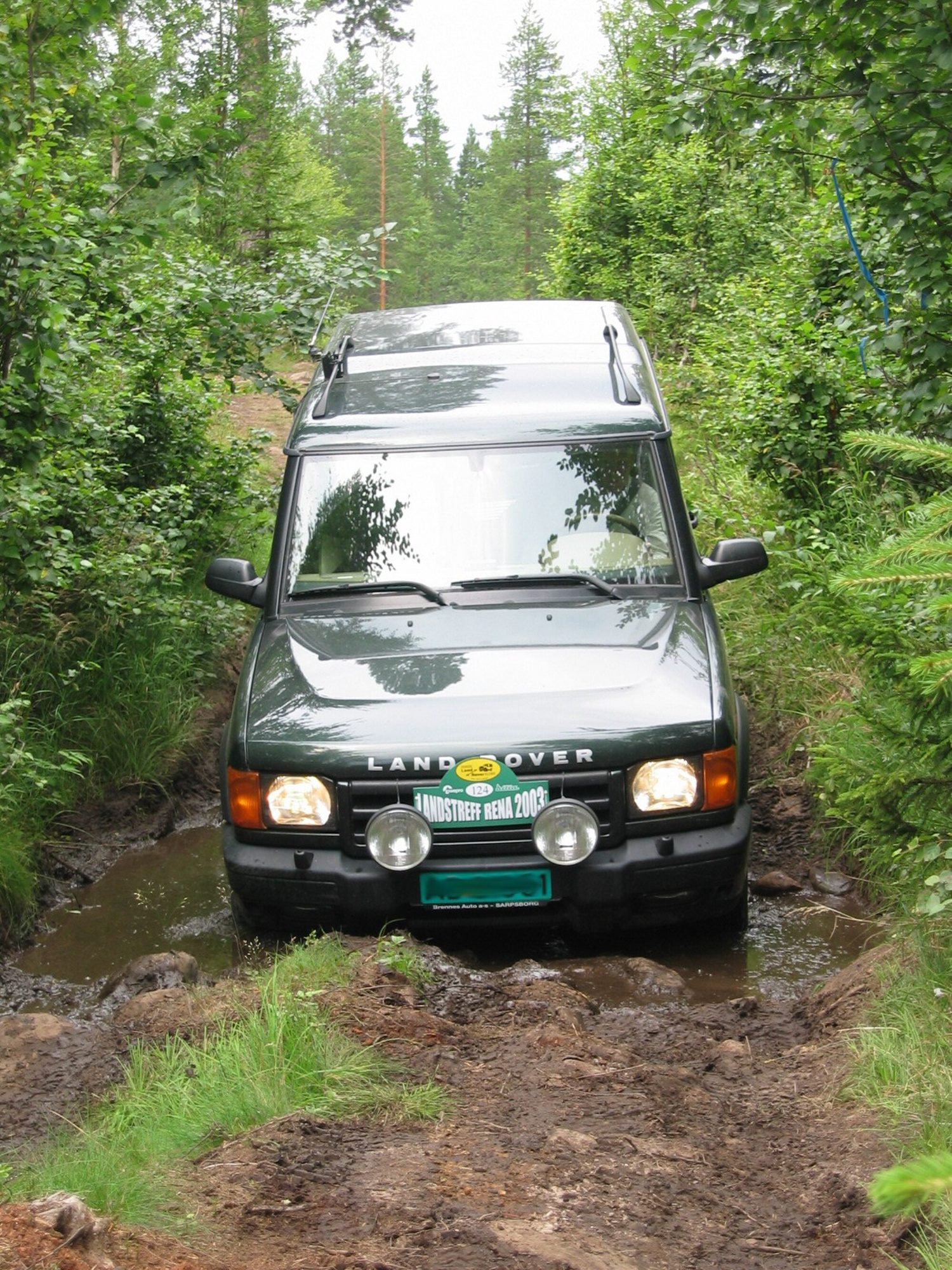 A dark green pre-facelift Discovery Series II with spotlights climbing out of a muddy forest track in Norway
