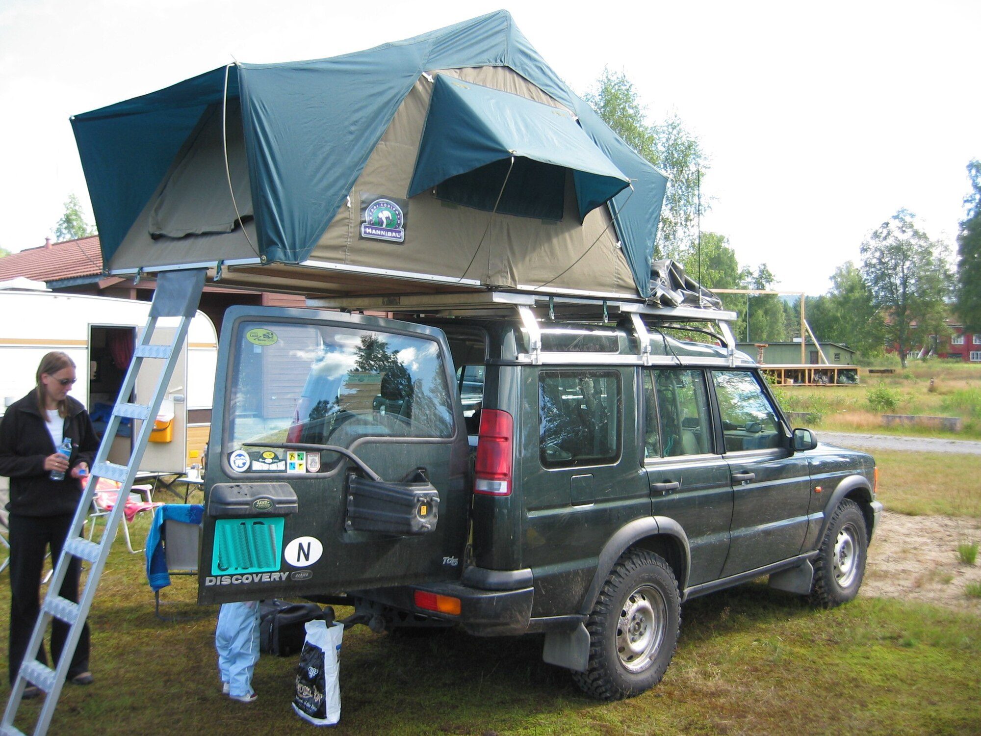 A dark green Discovery Series II Td5 with a Hannibal roof tent and ladder set up at a Land Rover club camp in a Norwegian field