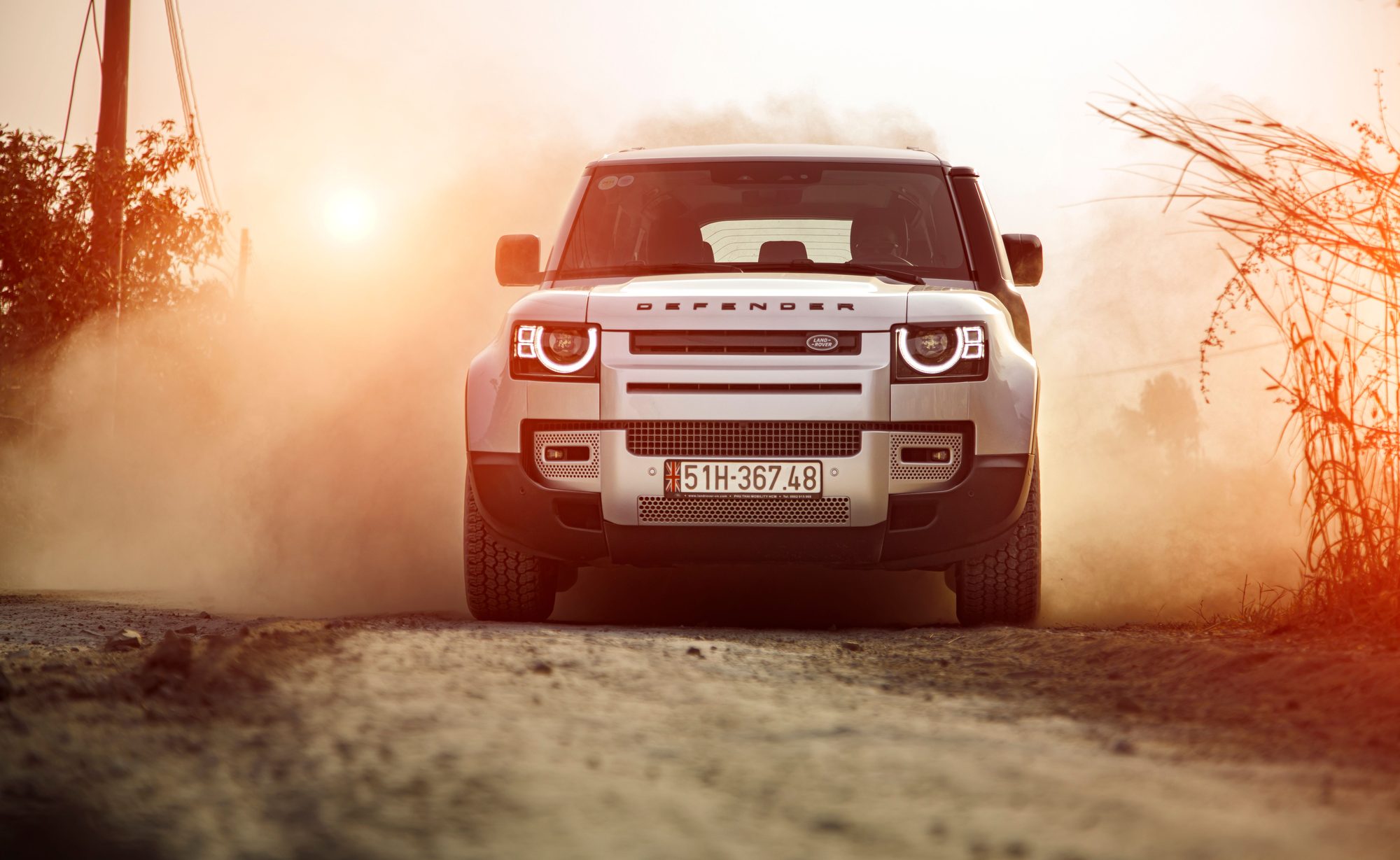 A silver Land Rover Defender (L663) driving head-on out of a cloud of dust on a dirt road at sunset