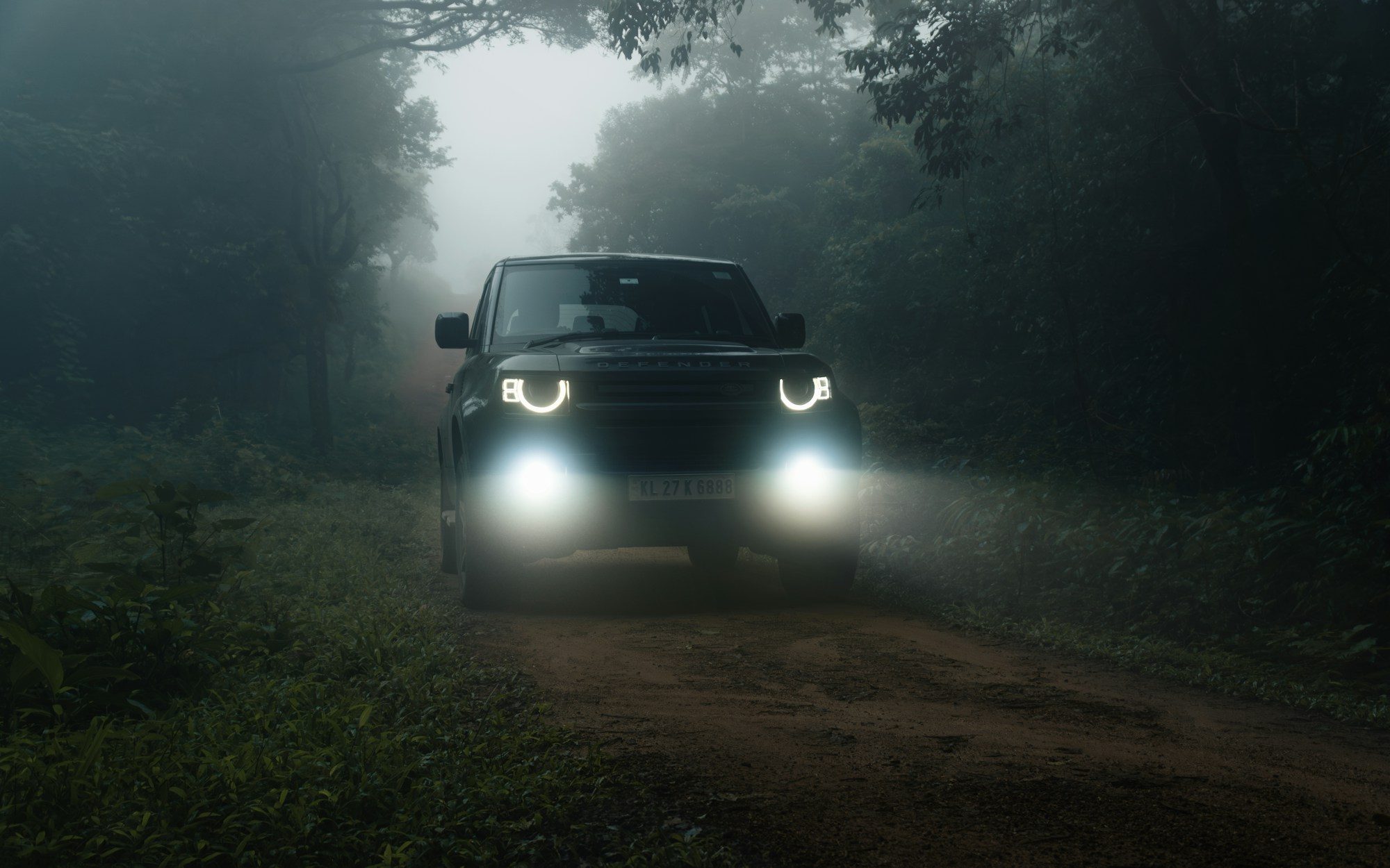 A dark Land Rover Defender (L663) with headlights and fog lamps on, on a red-earth forest track in mist, Idukki, Kerala