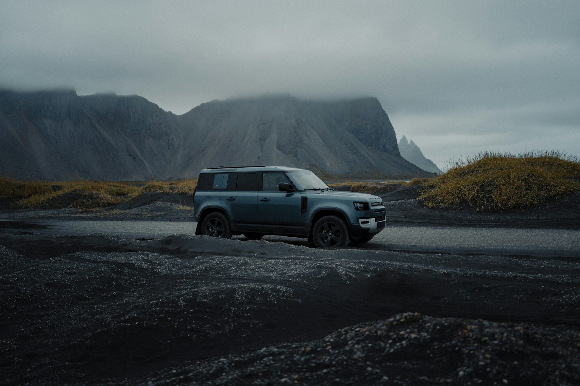 A blue-grey Land Rover Defender 110 (L663) on black volcanic sand below the cloud-capped Vestrahorn, Stokksnes, Iceland