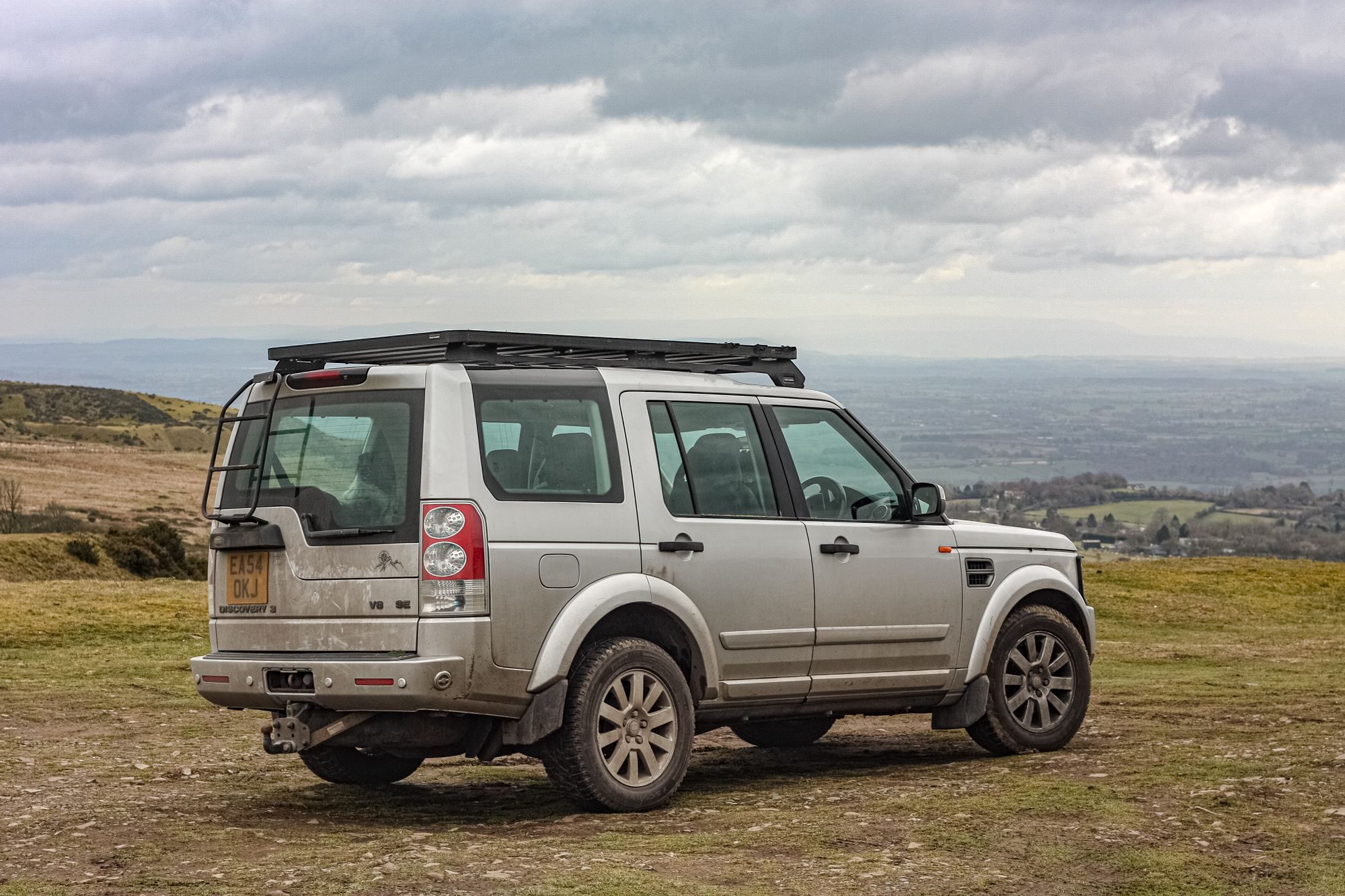 A silver Land Rover Discovery 3 V8 SE with a roof rack and rear ladder parked on rough grass on a hilltop overlooking a valley