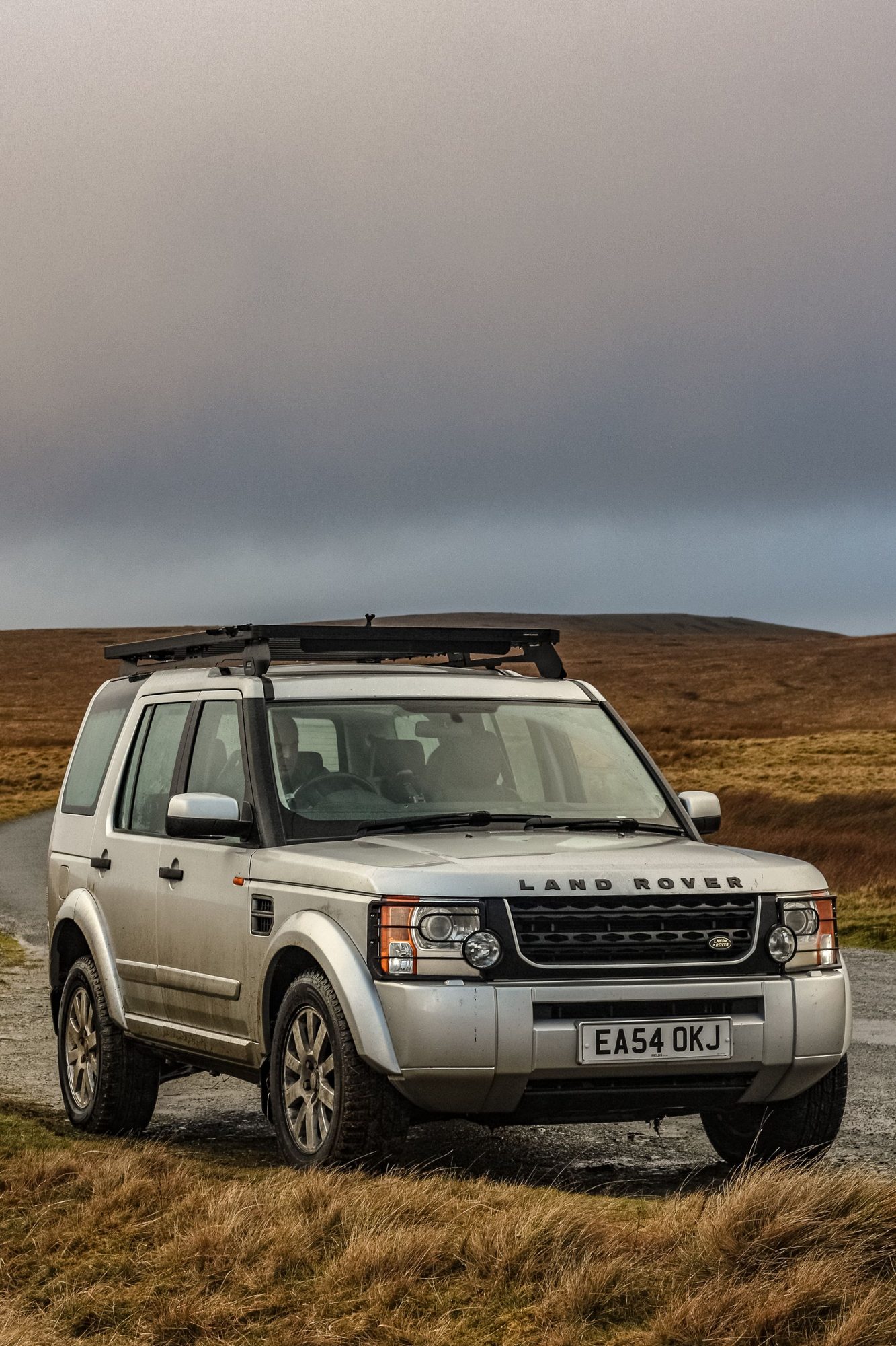 A silver Discovery 3 with a roof rack on a gravel moorland lane under a heavy grey sky
