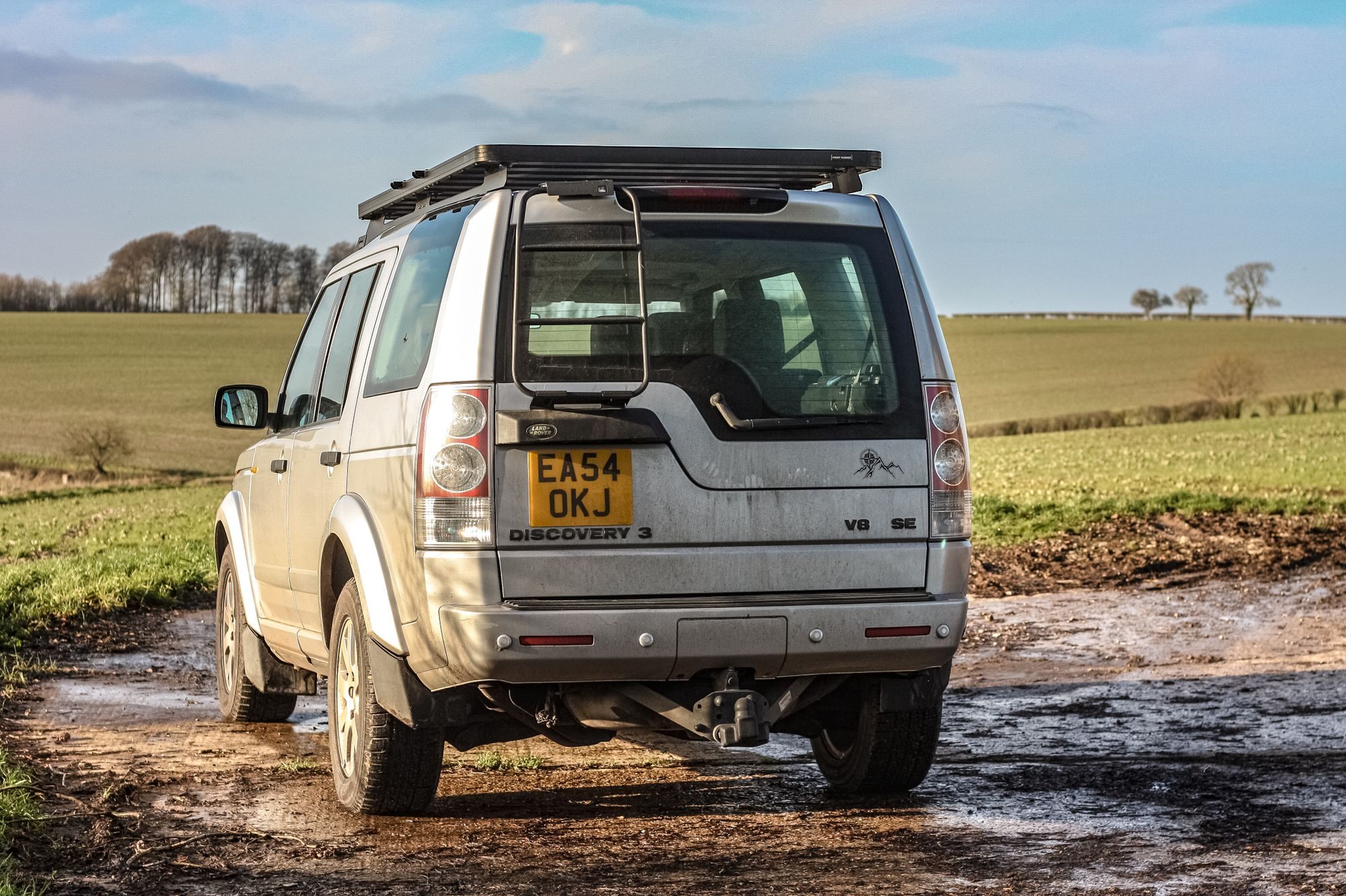 Rear view of a silver Discovery 3 V8 SE with roof rack and ladder on a muddy farm track between green fields