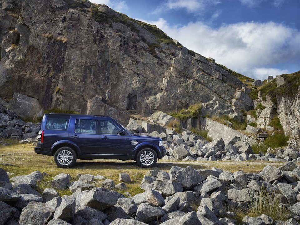 A dark blue 2014 facelift Discovery 4 parked on grass among boulders below a rock face