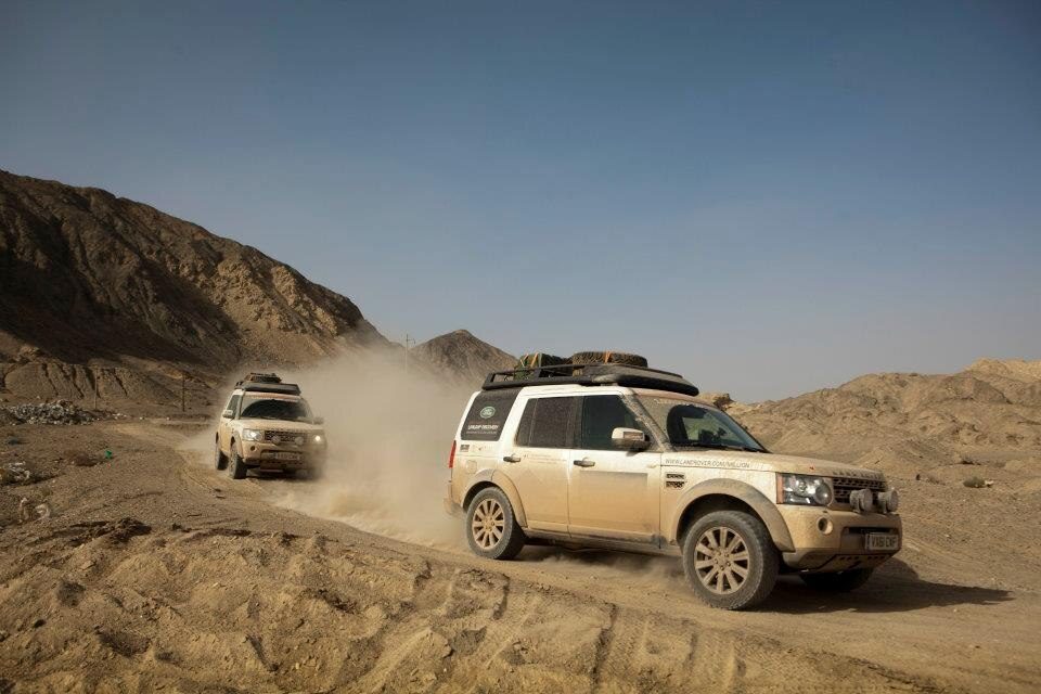 Two dusty Discovery 4s with roof racks and spare wheels kicking up dust on a desert track among bare hills in western China