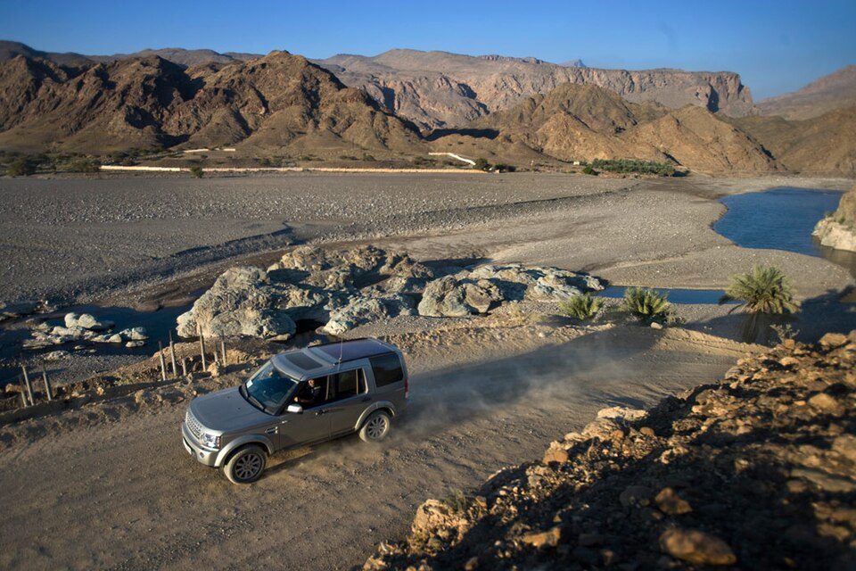 A silver Discovery 4 driving a dusty gravel track beside a wadi with palms and mountains in Oman