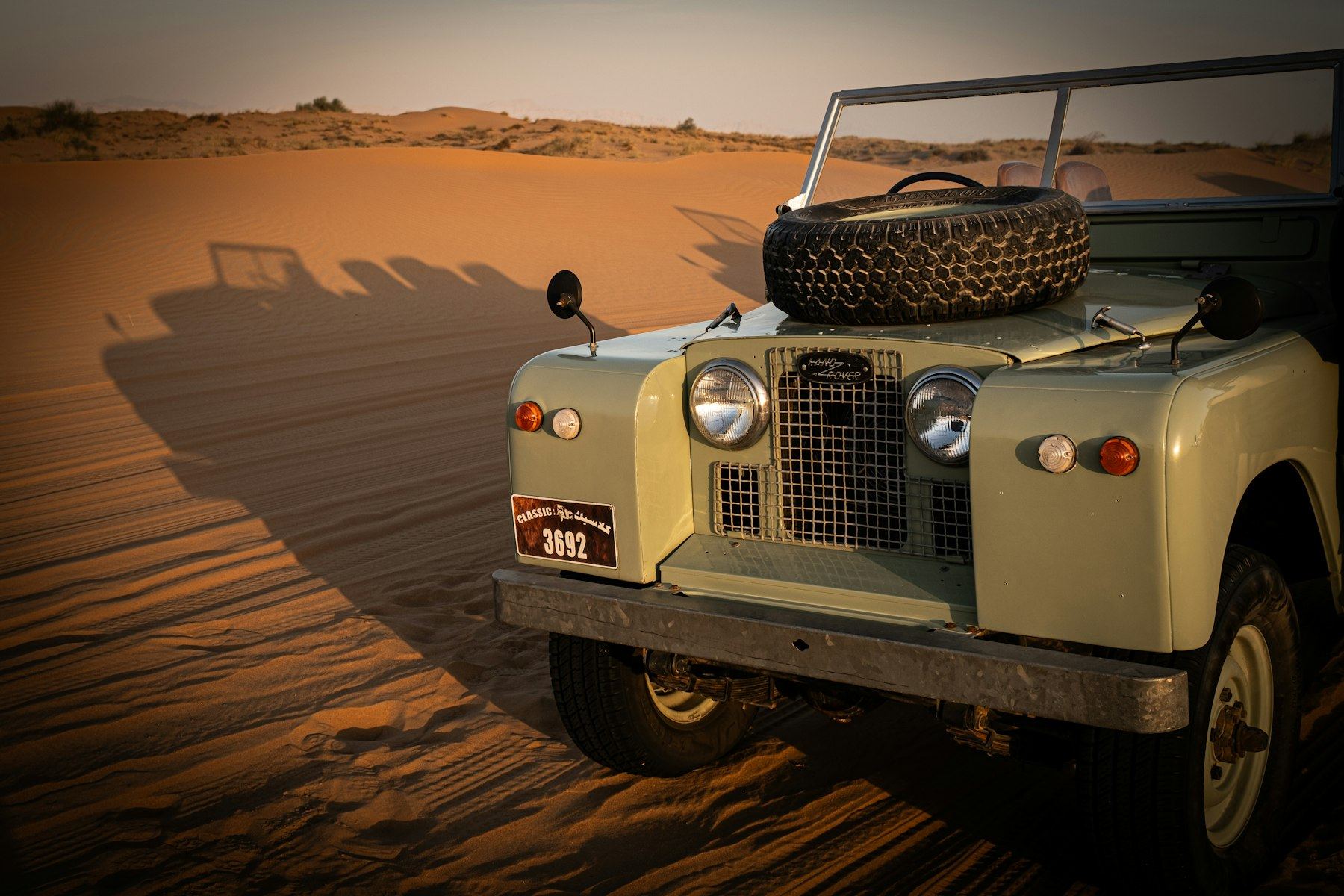 A sage-green Series II/IIA Land Rover with a bonnet-mounted spare wheel parked on desert sand at golden hour