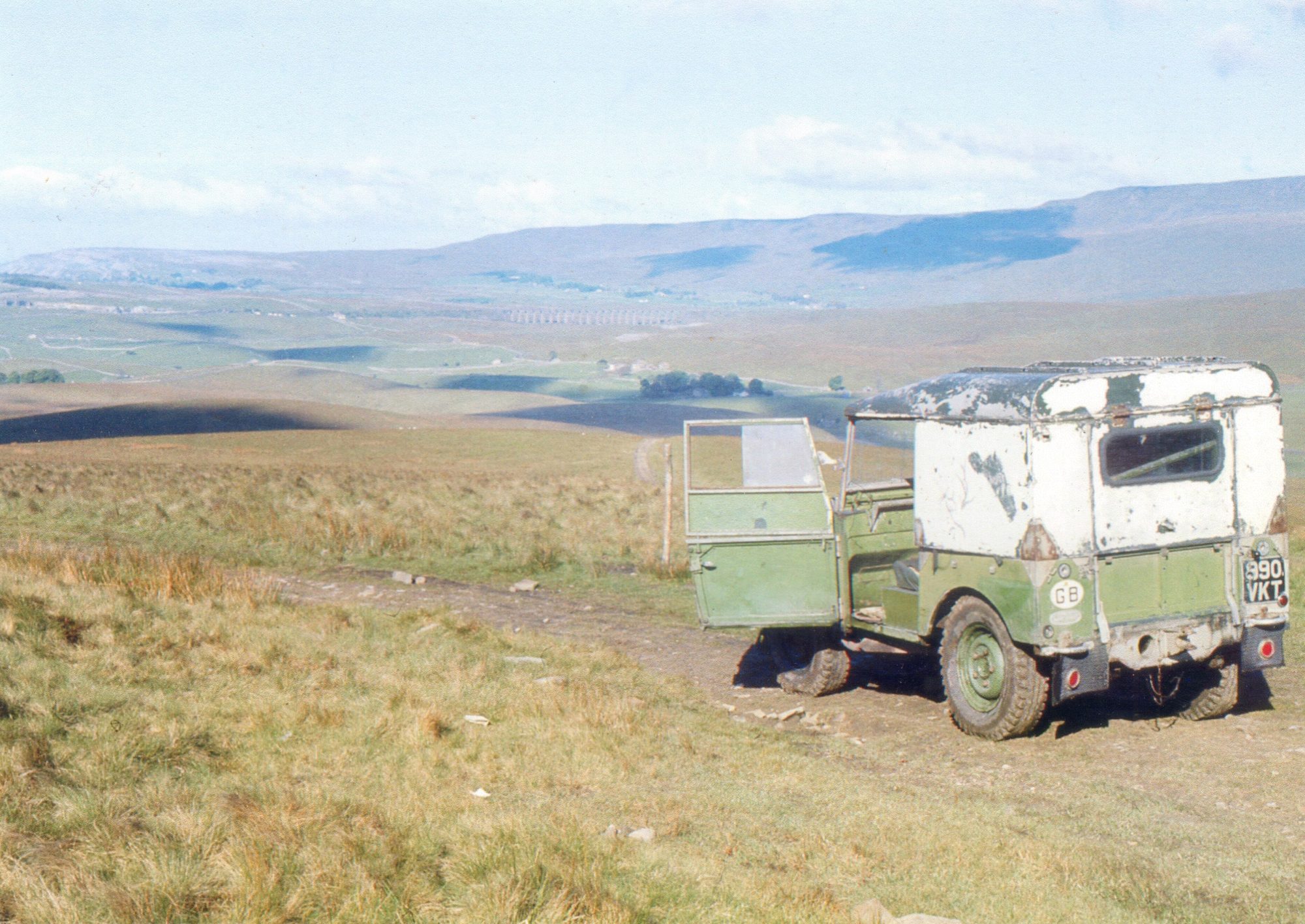 A weathered green Series I 80-inch Land Rover with a battered white hardtop on the Camm High Road moorland track, Ribblehead Viaduct in the valley below
