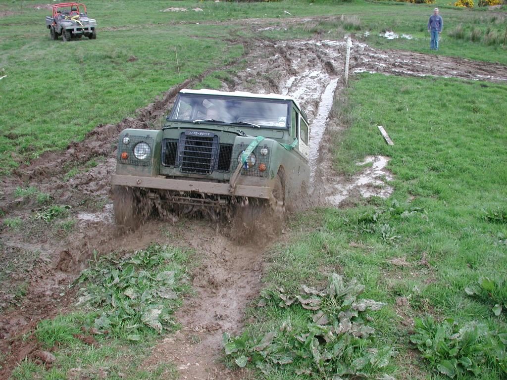 A green Series III Land Rover station wagon ploughing through a muddy rutted track in a field