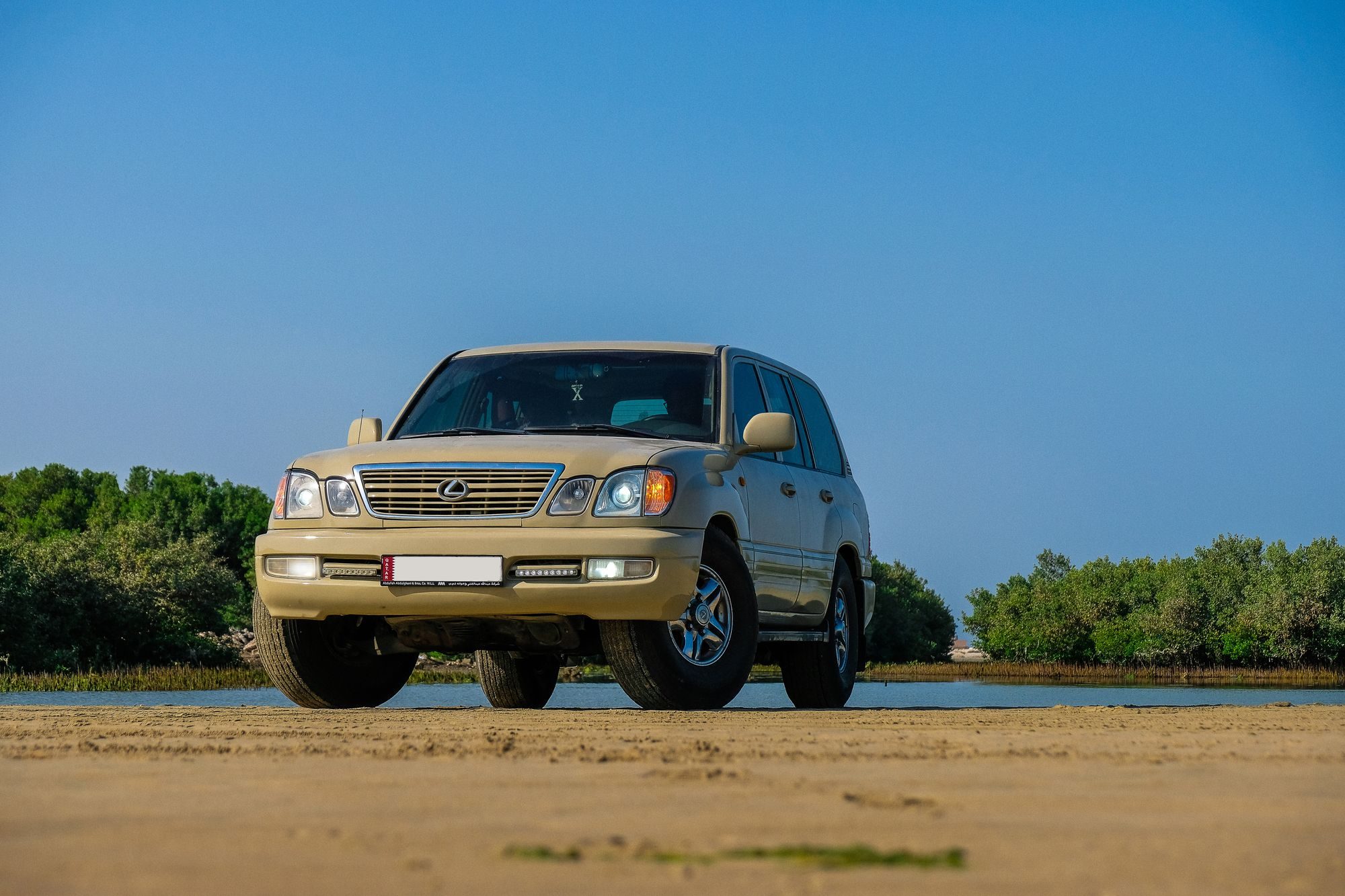 A low-angle view of a tan Lexus LX 470 on wet sand in front of a tidal inlet lined with mangroves
