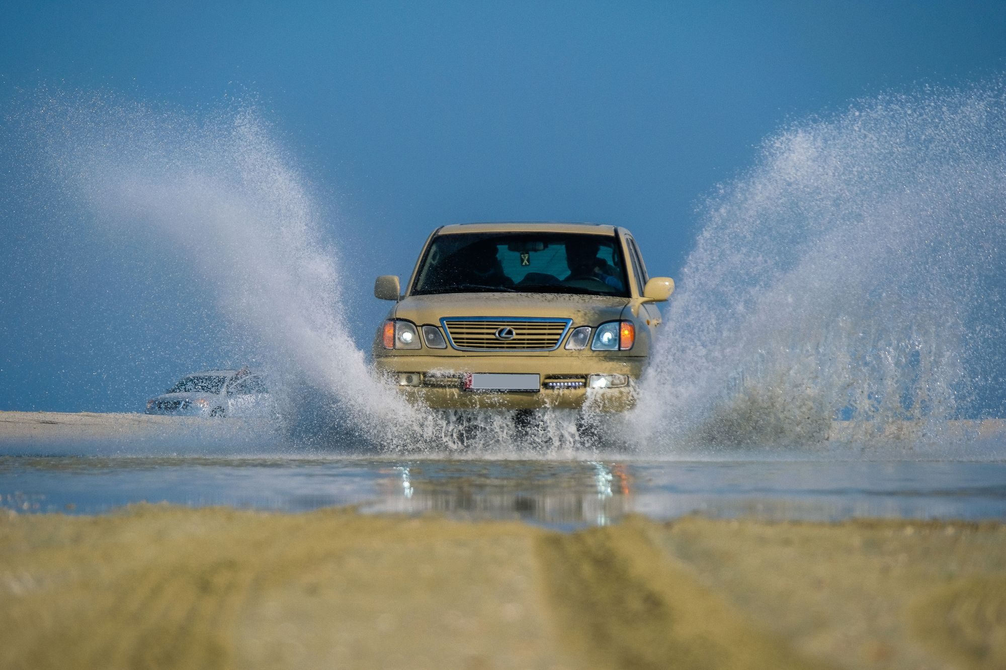 A tan Lexus LX 470 driving head-on through standing water on sand flats, throwing up two walls of spray