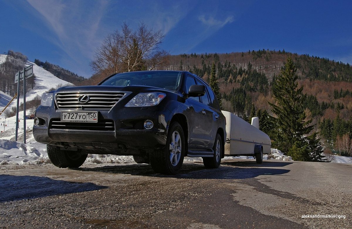 A black early Lexus LX 570 towing a glider trailer on a snowy mountain pass