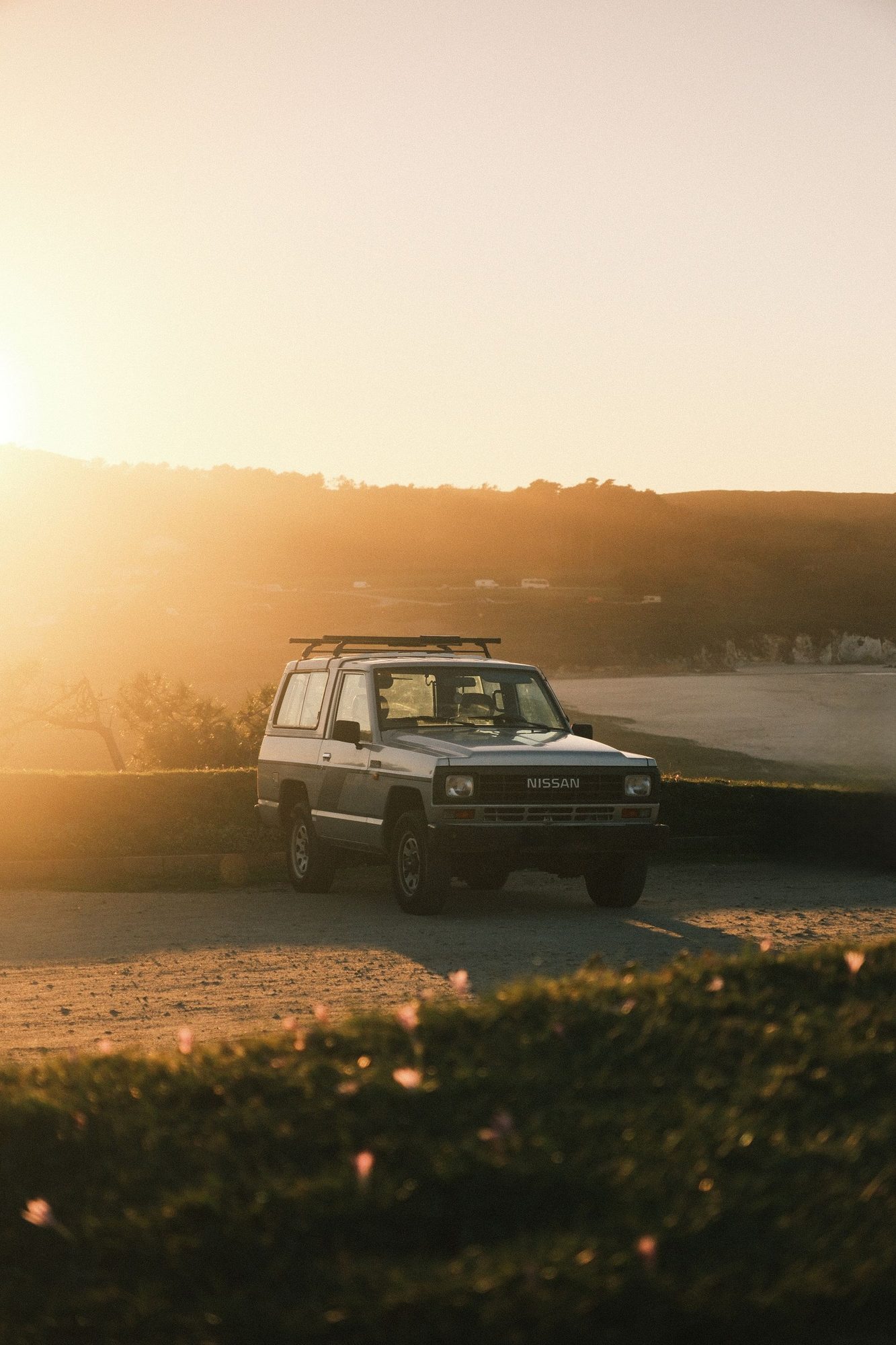 A silver two-door 160-series Nissan Patrol with rectangular headlights and a roof rack parked on a gravel clifftop above a beach at sunset