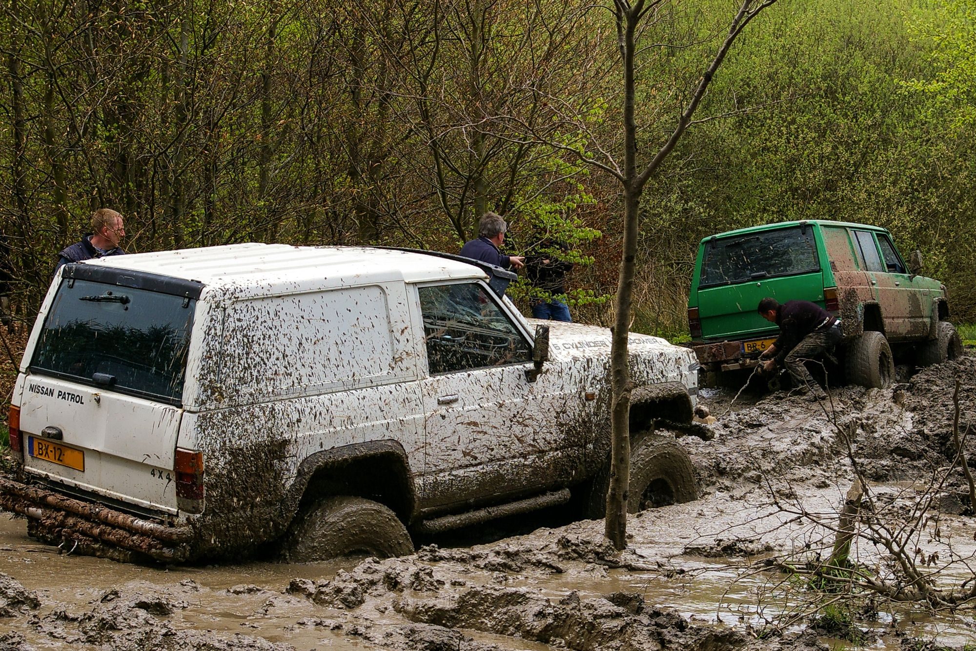 A mud-spattered white short-wheelbase 160-series Nissan Patrol hardtop sunk to its axles in a flooded woodland track, with a green long-wheelbase 160 being winched out ahead