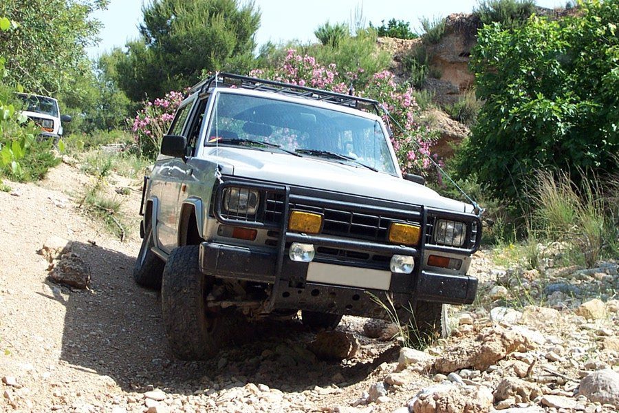 A silver short-wheelbase leaf-sprung Nissan Patrol with rectangular headlights, a bull bar and roof rack, climbing a rocky track between bushes
