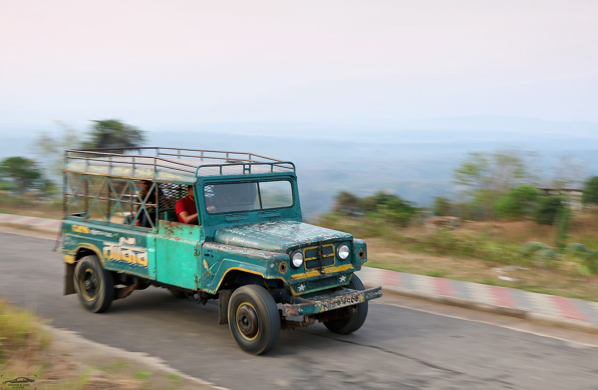 A teal Nissan Patrol 60 converted into a Chander Gari shared taxi, with a roof cage, driving along a hill road above a hazy valley in Bangladesh