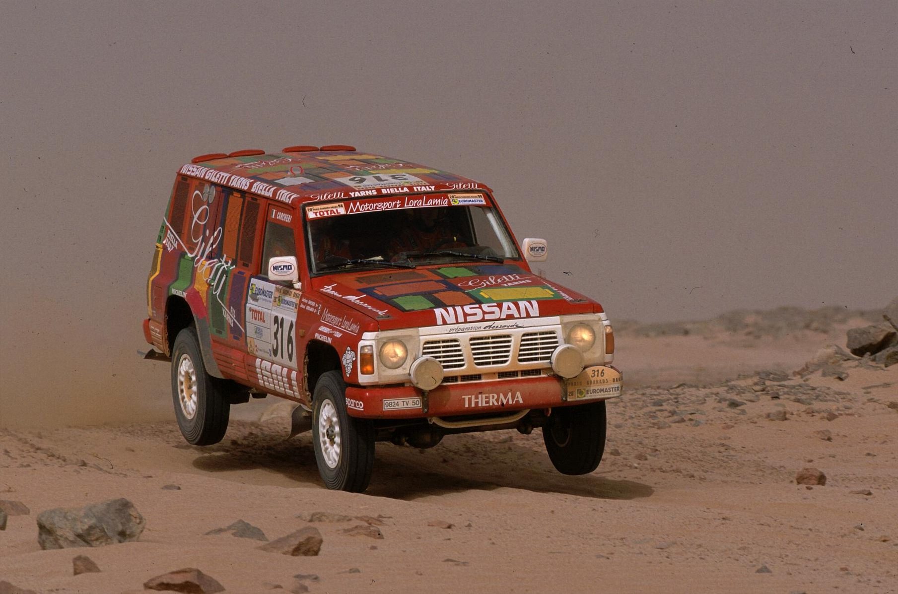 A red Team Nissan France Dessoude Patrol GR Y60 4.2 T1 airborne on a rocky desert stage of the 1998 Paris–Granada–Dakar rally
