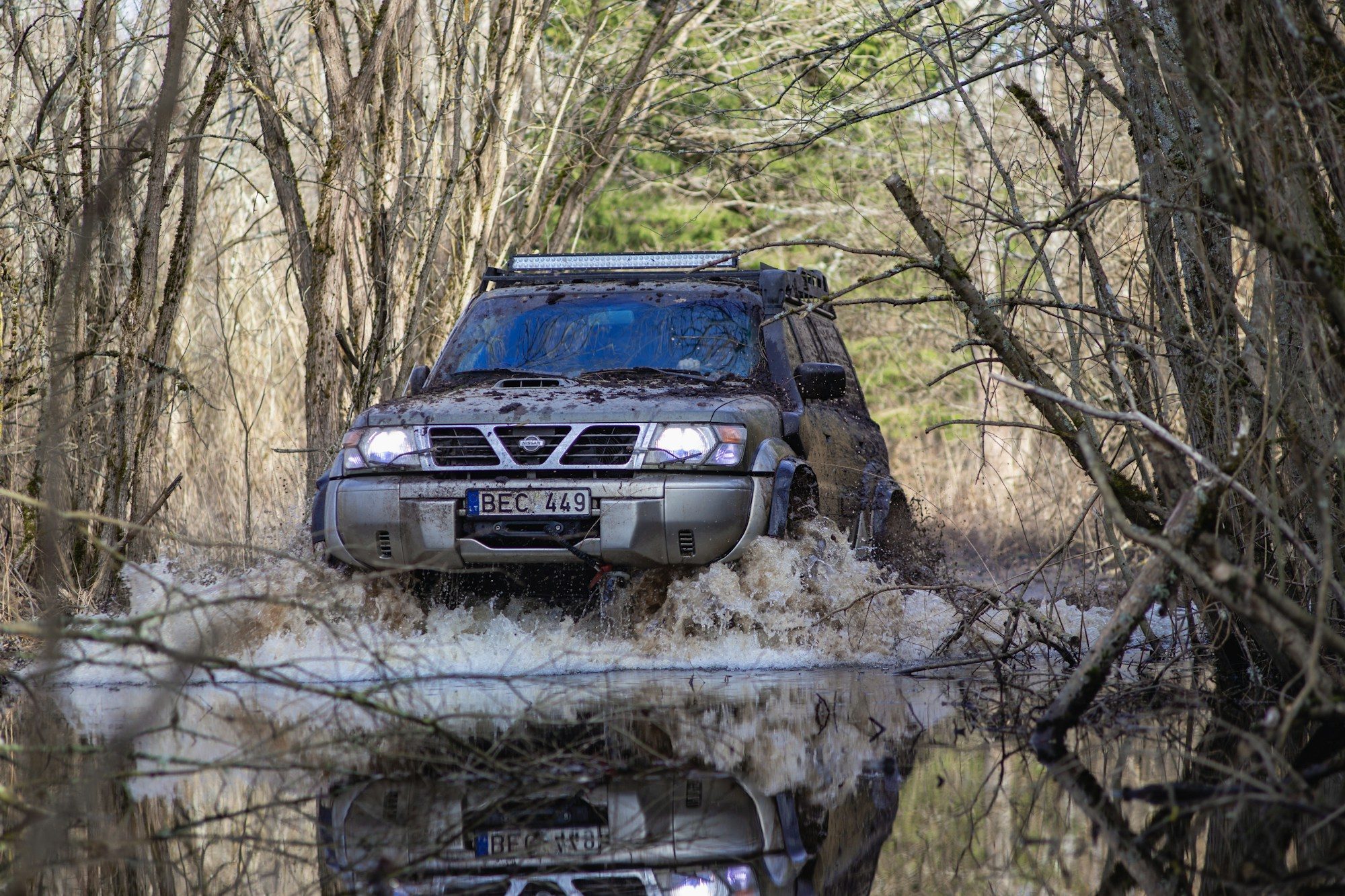 A muddy pre-facelift Nissan Patrol Y61 with a snorkel, winch and roof-mounted light bar ploughing through a flooded forest track in Lithuania