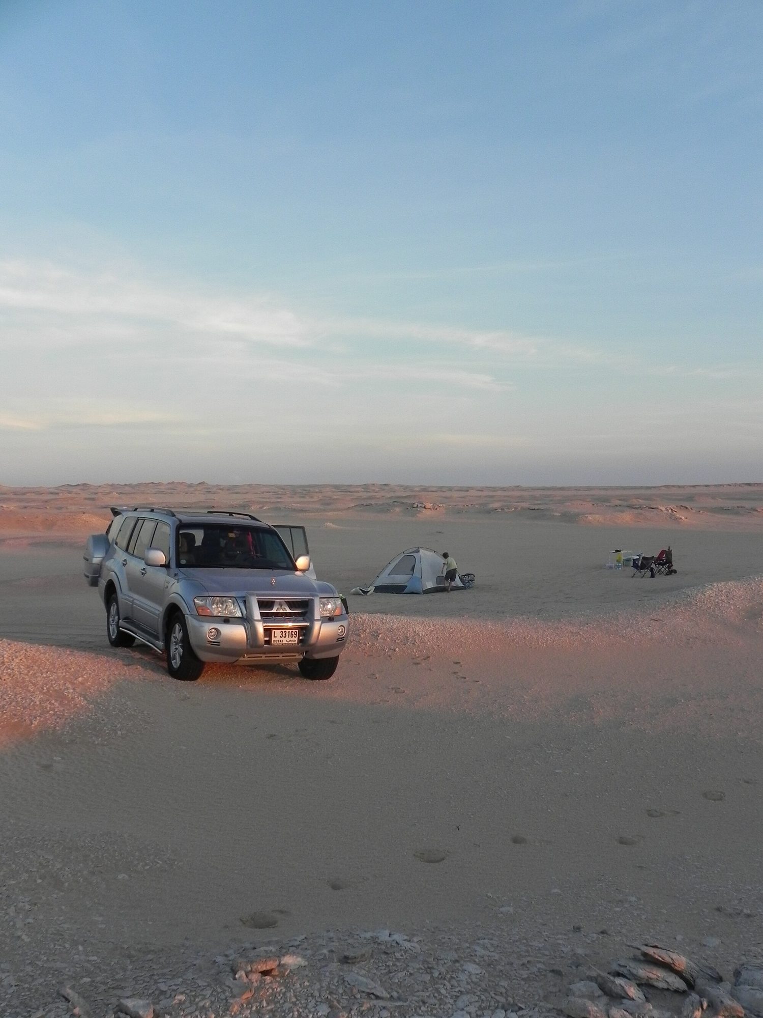 A silver facelifted third-generation Pajero with a bull bar parked at a desert campsite at sunset, a tent pitched beside it