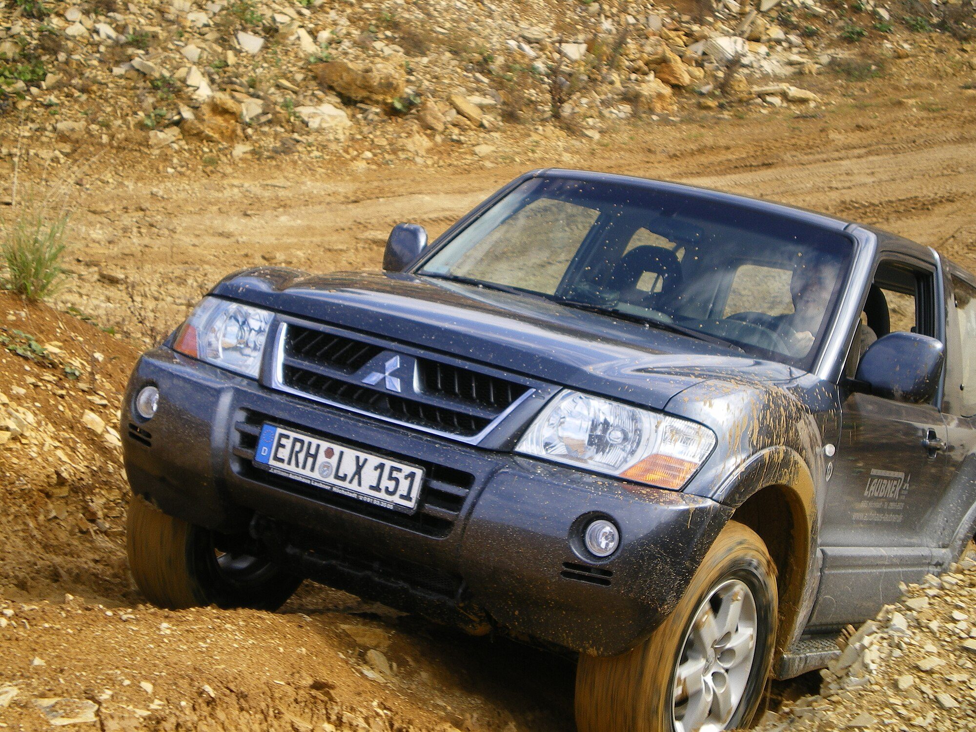 A mud-spattered dark blue facelifted third-generation Pajero cross-axled on a dirt and rock slope