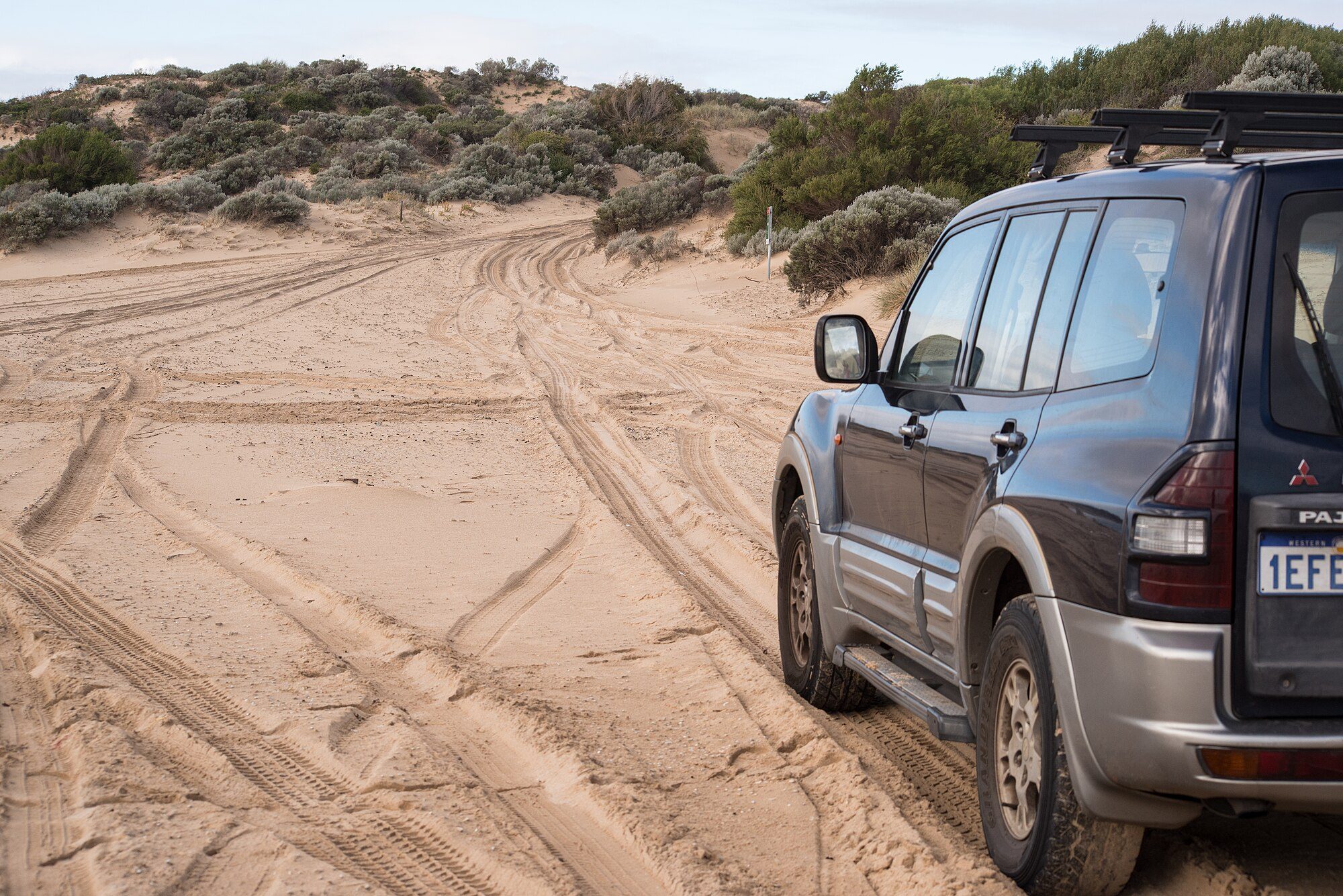 A dark grey and silver pre-facelift third-generation Pajero with roof bars on a rutted sand track through coastal dunes at Tim's Thicket, Western Australia