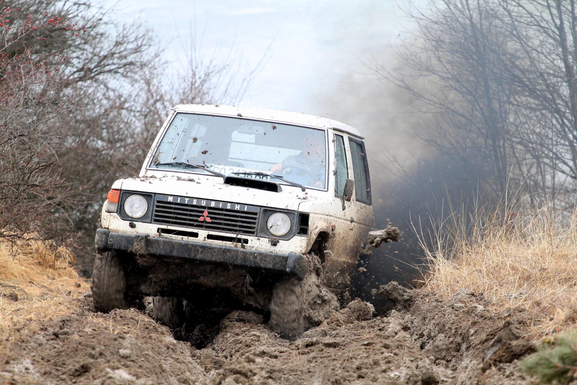 A white first-generation Pajero three-door ploughing through deep mud with dirt flying off its wheels at a Bulgarian off-road event