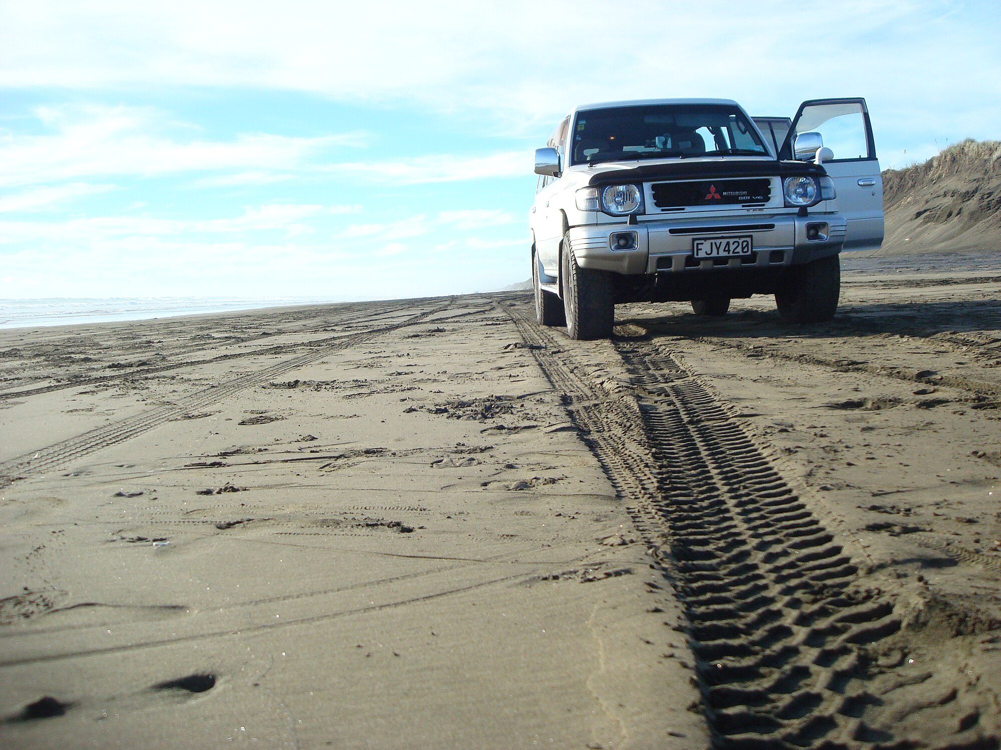 A silver late second-generation Pajero GDI V6 with its door open on the black-sand 4x4 track at Muriwai Beach, New Zealand