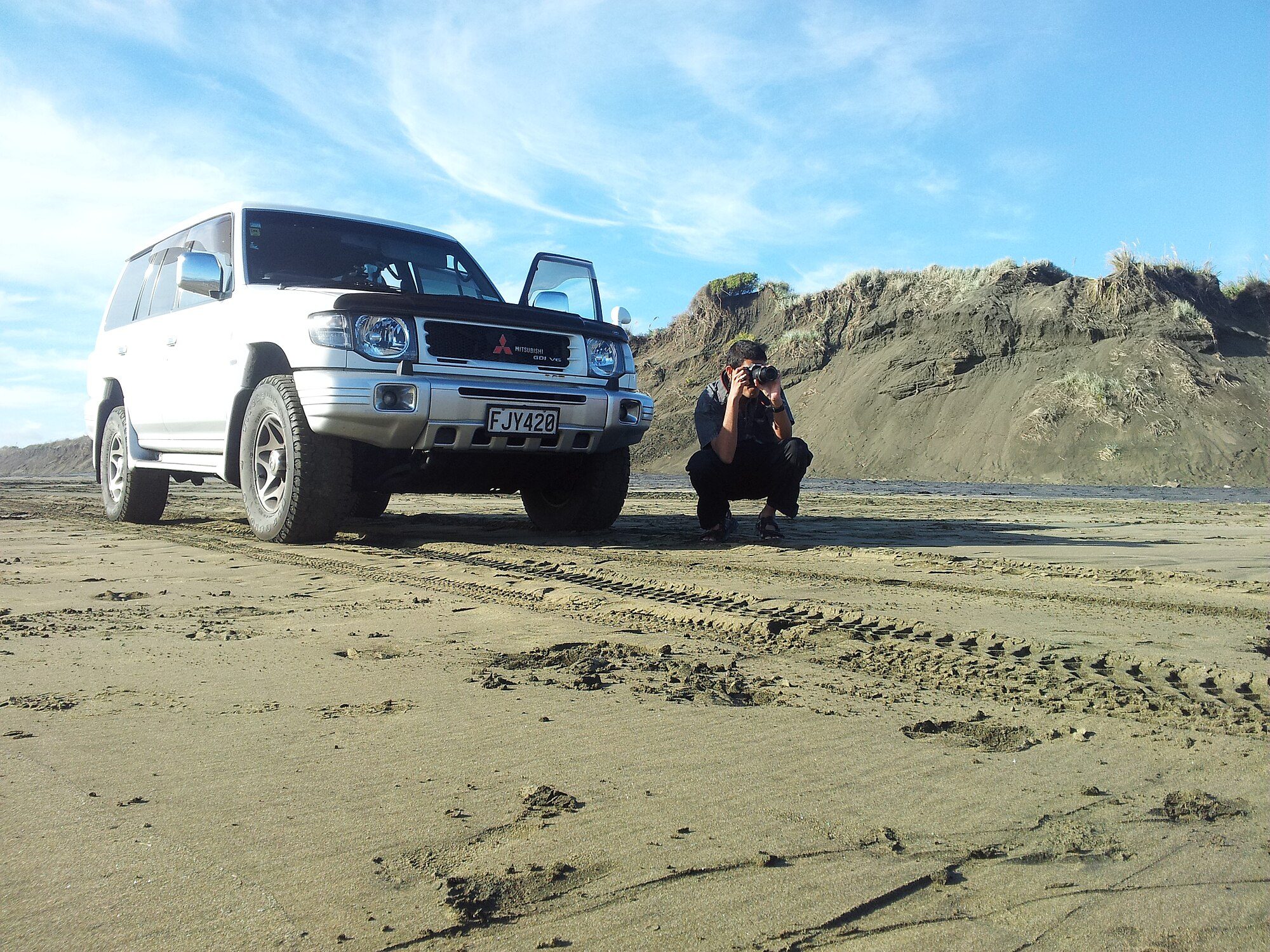 A white late second-generation Pajero parked on the sand below grassy dunes on the Muriwai 4x4 track while a photographer crouches beside it