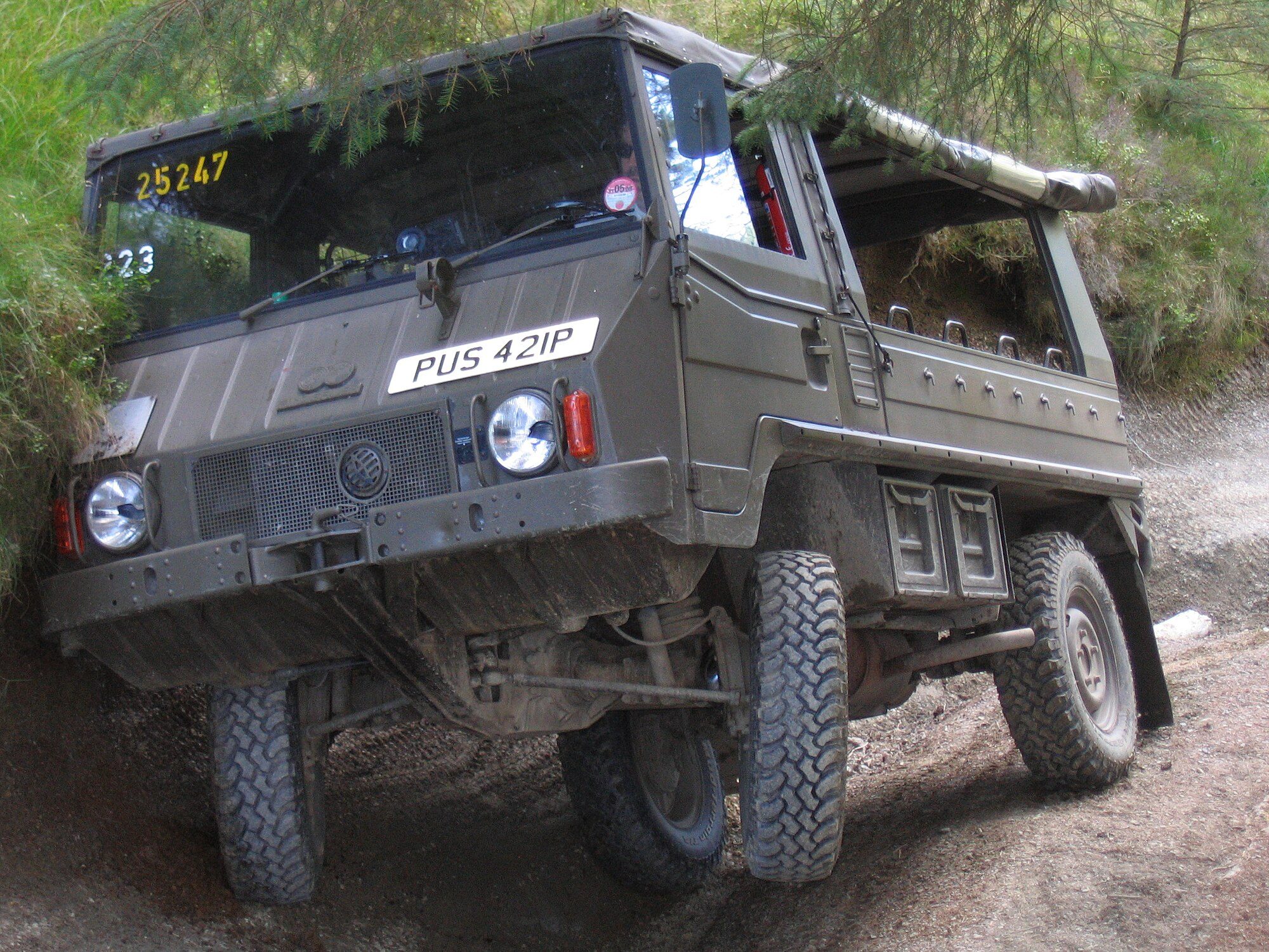 An olive-green ex-Swiss Army Pinzgauer 710 soft top flexing its swing axles on a muddy wooded byway in Wales