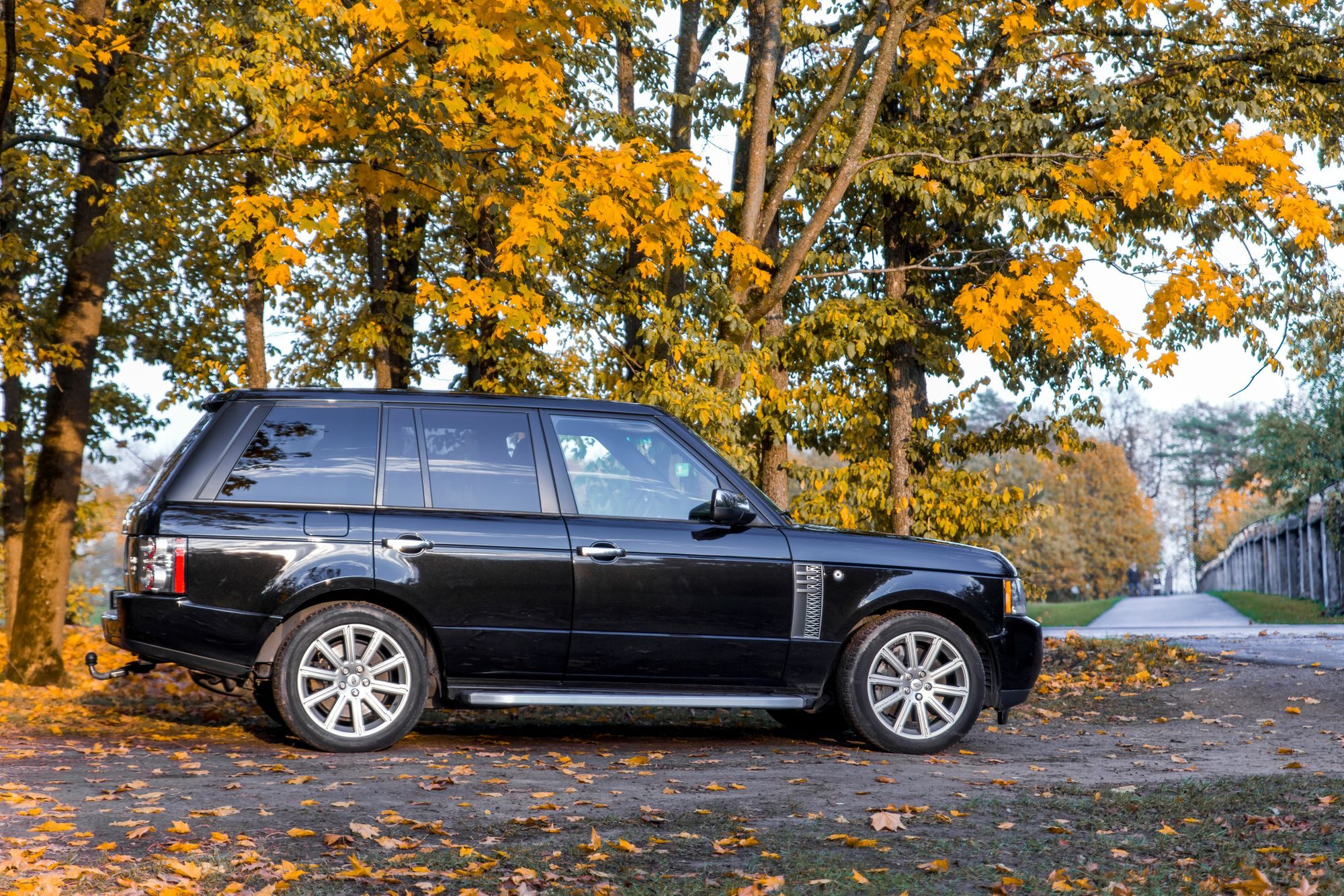 A black facelifted L322 Range Rover in profile on a leaf-covered gravel clearing among autumn trees
