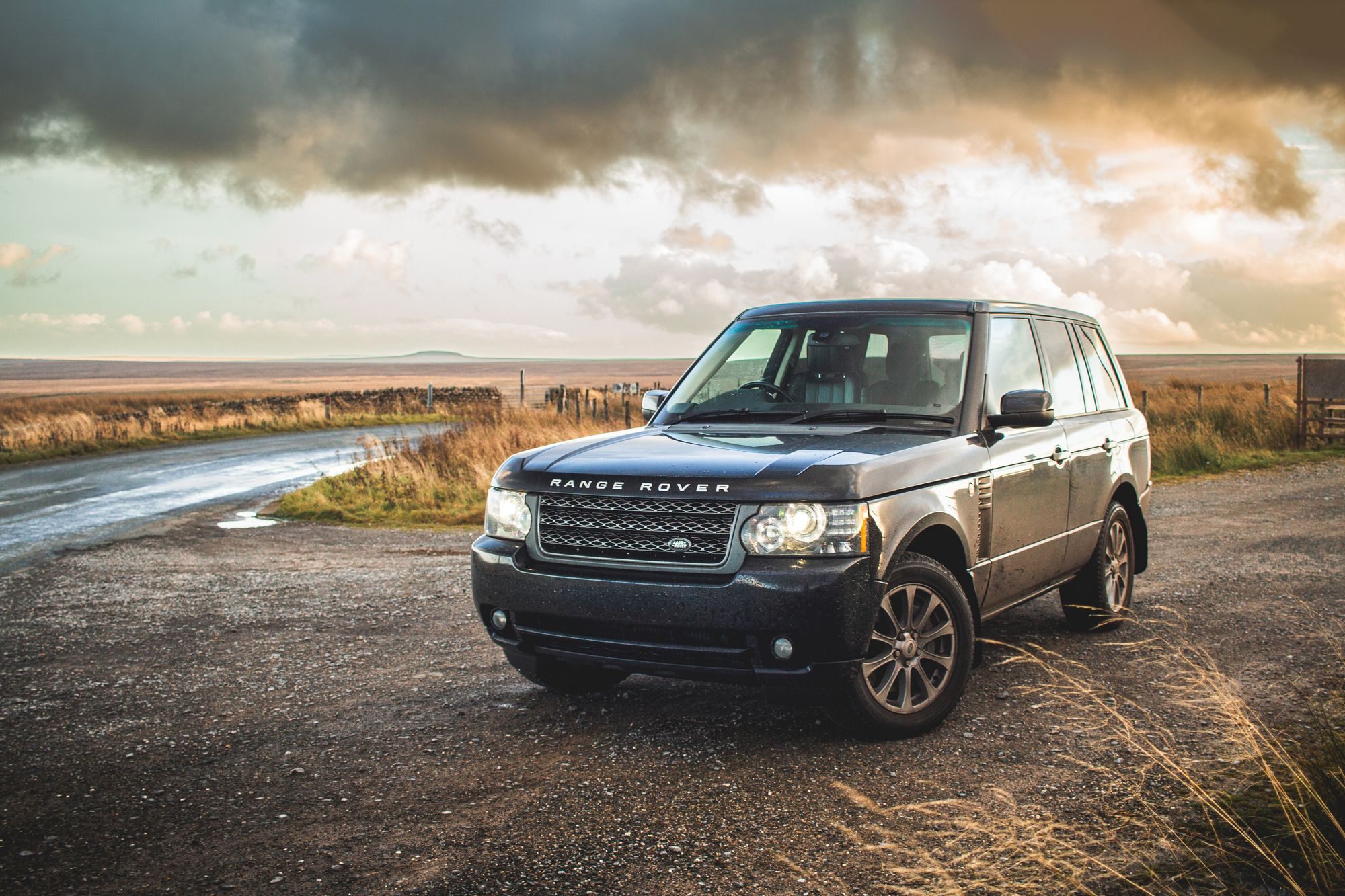 A dark facelifted third-generation Range Rover (L322) on a gravel pull-off beside a moorland lane under storm clouds