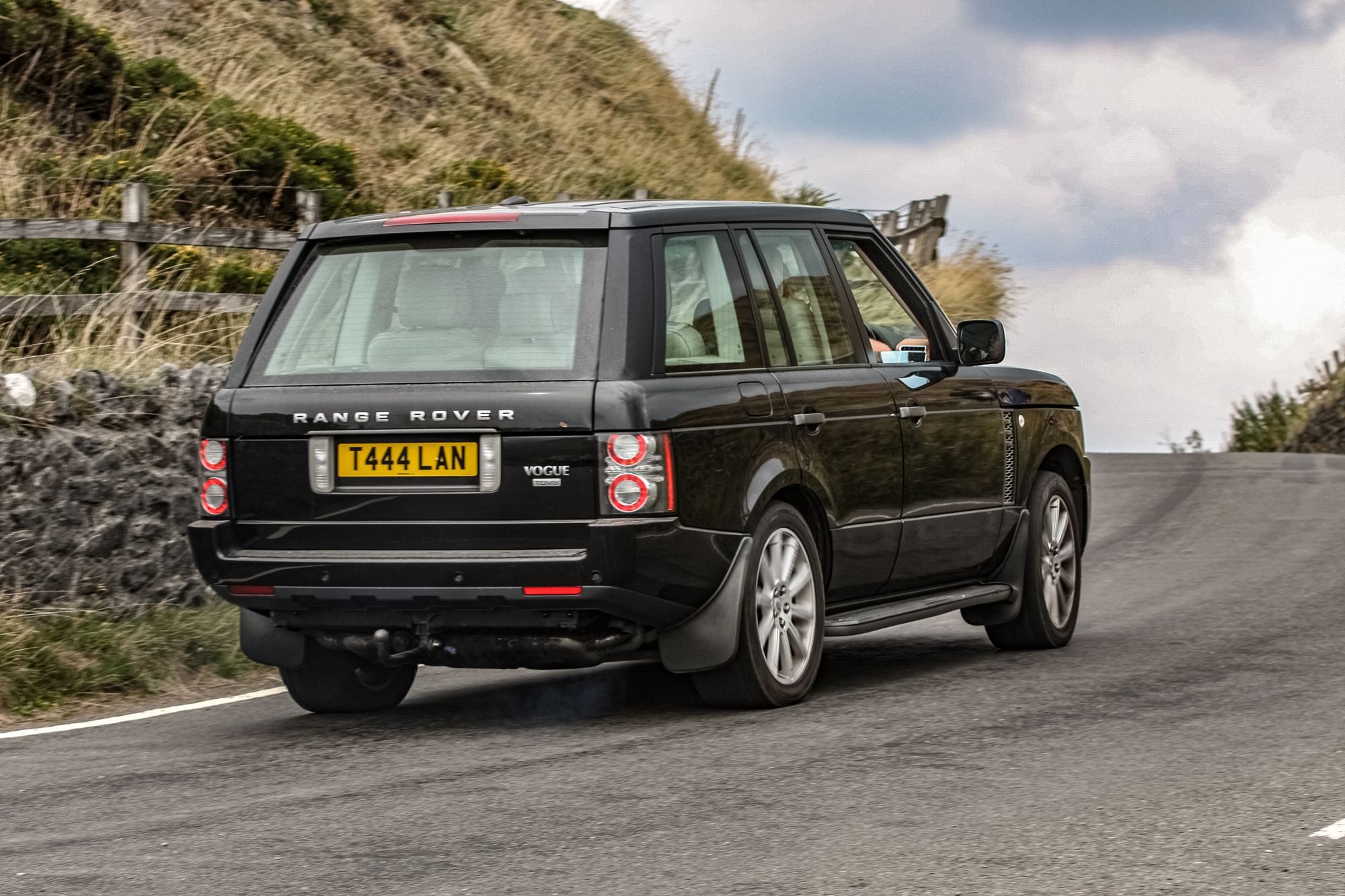 A black L322 Range Rover Vogue seen from behind climbing a narrow hill road past a dry-stone wall